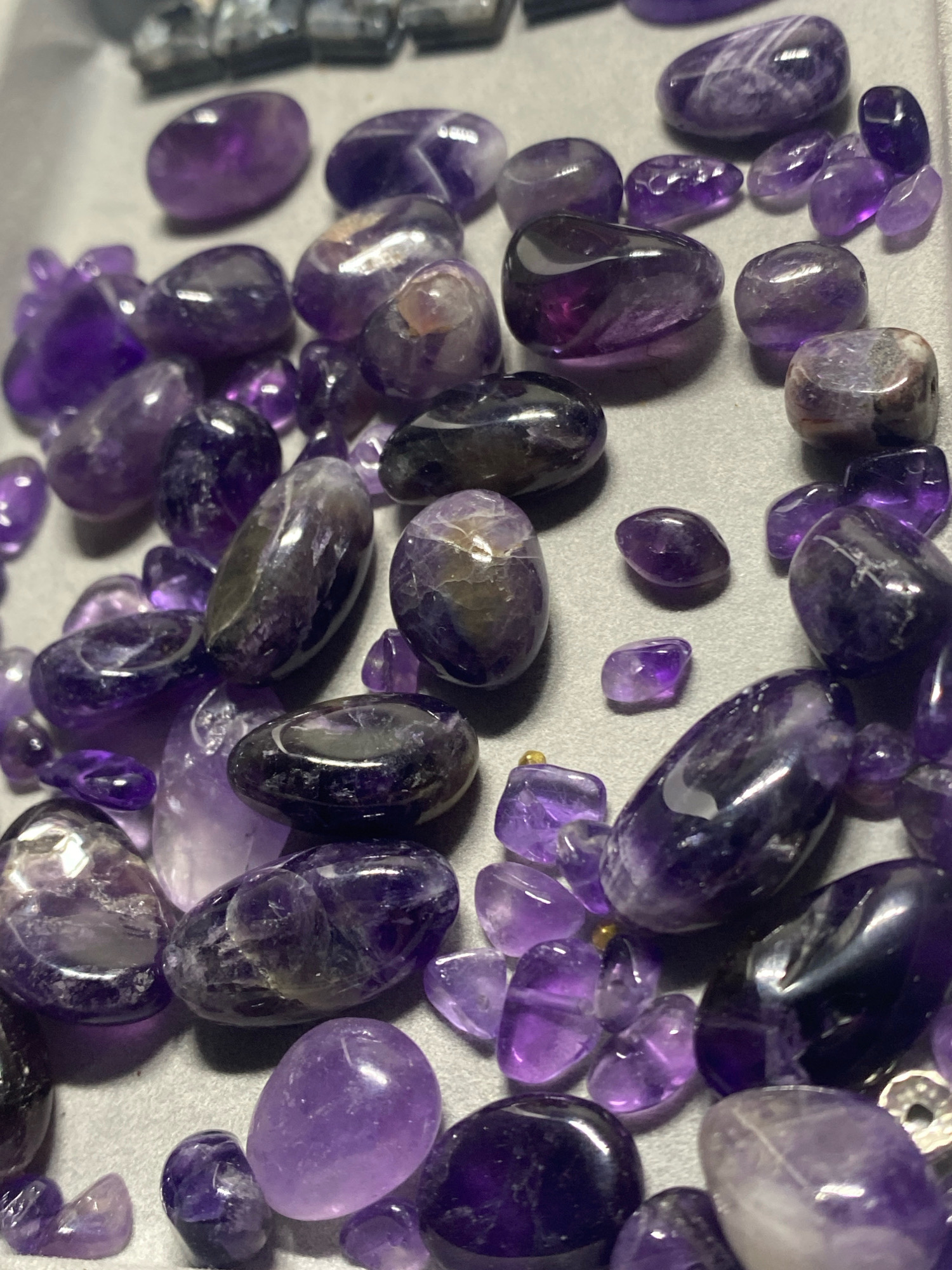 The image displays a close-up, overhead view of numerous tumbled amethyst stones scattered on a neutral gray surface. The stones vary in size, shape, and shade of purple, ranging from deep, translucent violet to lighter, milky lavender. Some stones exhibit internal banding and inclusions, creating a varied texture and visual appeal. There are no people, animals, or discernable landmarks present. The scene is static and appears to be a collection of gemstones. The location of Kushchovskaya, Russia, is not depicted or suggested by any visual cues in the image.