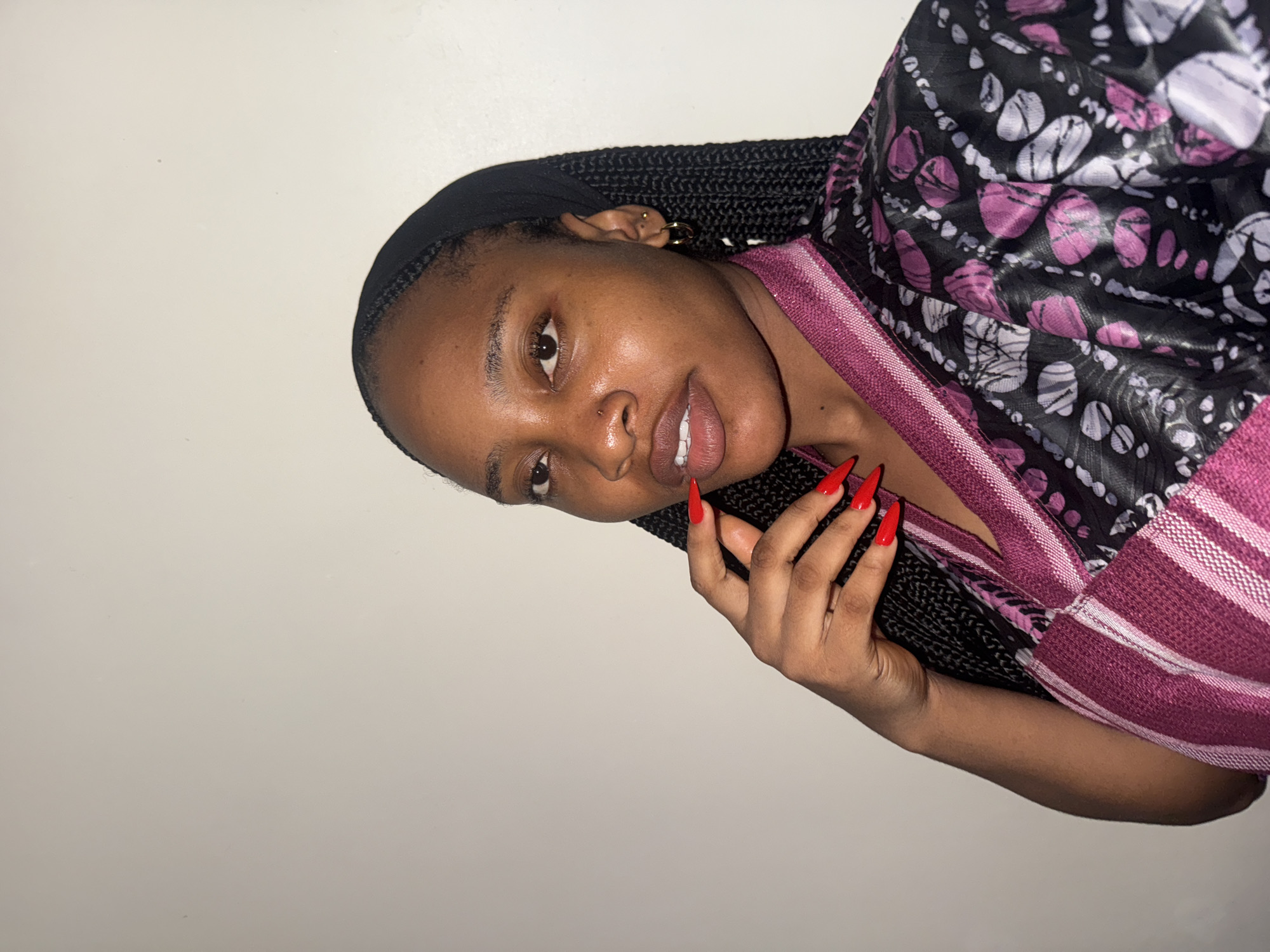A young Black woman is depicted in a close-up portrait, oriented diagonally in the frame, against a plain, light-colored wall. She looks directly at the camera with a calm expression and a slight smile.
Her hair is styled in long, thin black braids pulled back from her face. She accessorizes with a gold hoop earring in her right ear, a smaller gold stud in her left, and a subtle stud piercing on her right nostril. Her skin appears smooth and well-lit.
She is wearing a top or dress with a striking pattern: a dark background featuring irregular, abstract shapes in shades of white and purple/magenta. A vibrant, solid magenta fabric, possibly textured or woven with subtle stripes, forms a trim or border on her garment, visible around her upper body. Her left hand is raised near her chin, prominently displaying long, pointed (stiletto) nails painted a bright red.
The setting appears to be indoors, indicated by the uniform, unadorned light background. There is no visible text in the image. The overall impression is a direct and composed portrait, consistent with an individual from Uyo, Nigeria.
