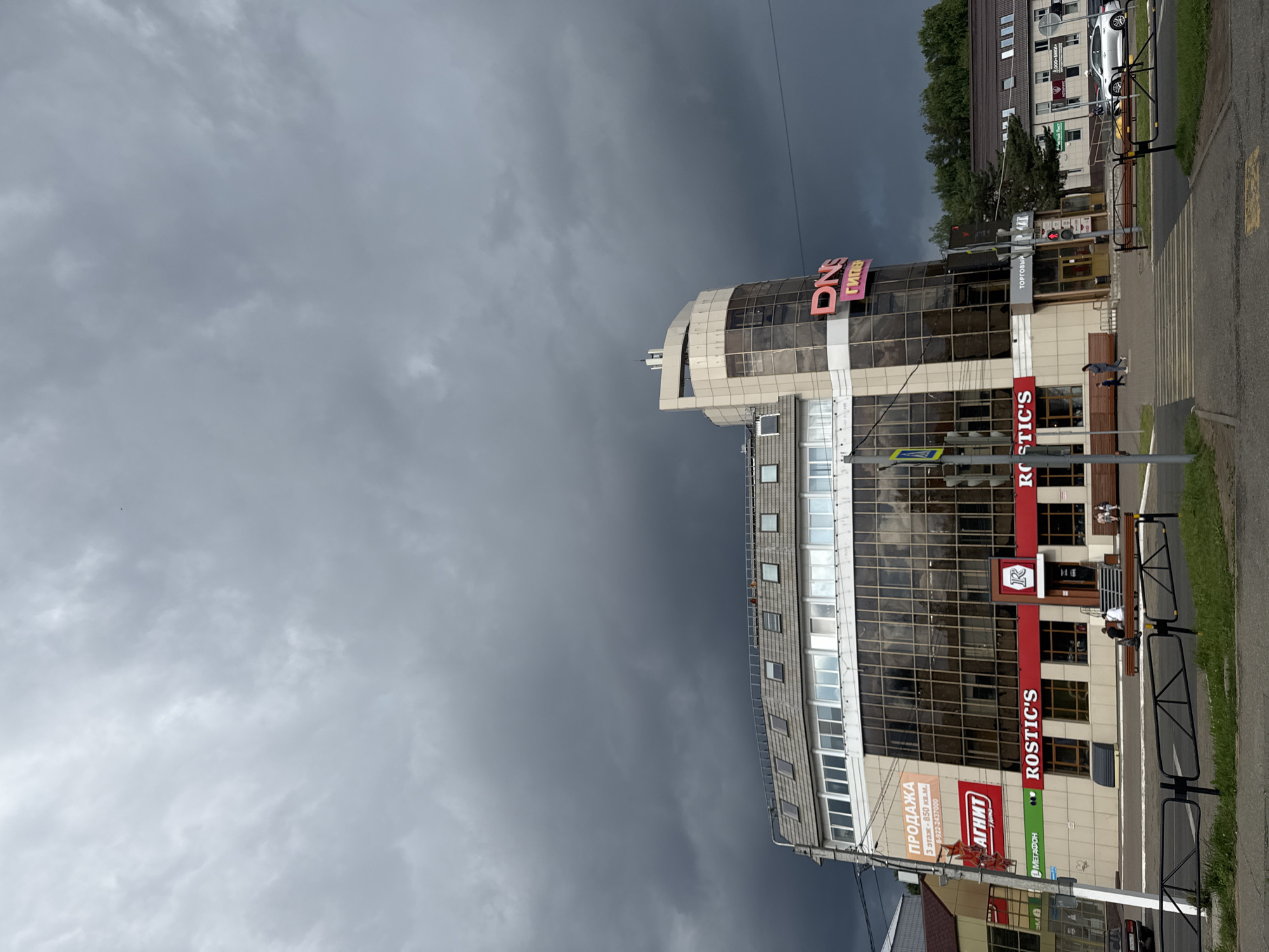 The image shows a commercial building in the city of Chaikovsky, Russia, on a cloudy day. The building has a modern design with large windows and several visible storefronts. Notable signs include "DNS" in large pink letters and "ROSTIC'S" in red letters. There are also signs for "ПРОДАЖА" (Sale) and "АГНИТ" (Agniť), which appears to be a supermarket chain. The ground floor of the building features multiple businesses. In front of the building, there is a sidewalk with benches, a crosswalk, and a traffic light. A few people are visible walking on the sidewalk and near the crosswalk. The sky is filled with dark, ominous clouds, suggesting an impending storm. The overall scene depicts a typical urban commercial area under a dramatic sky.