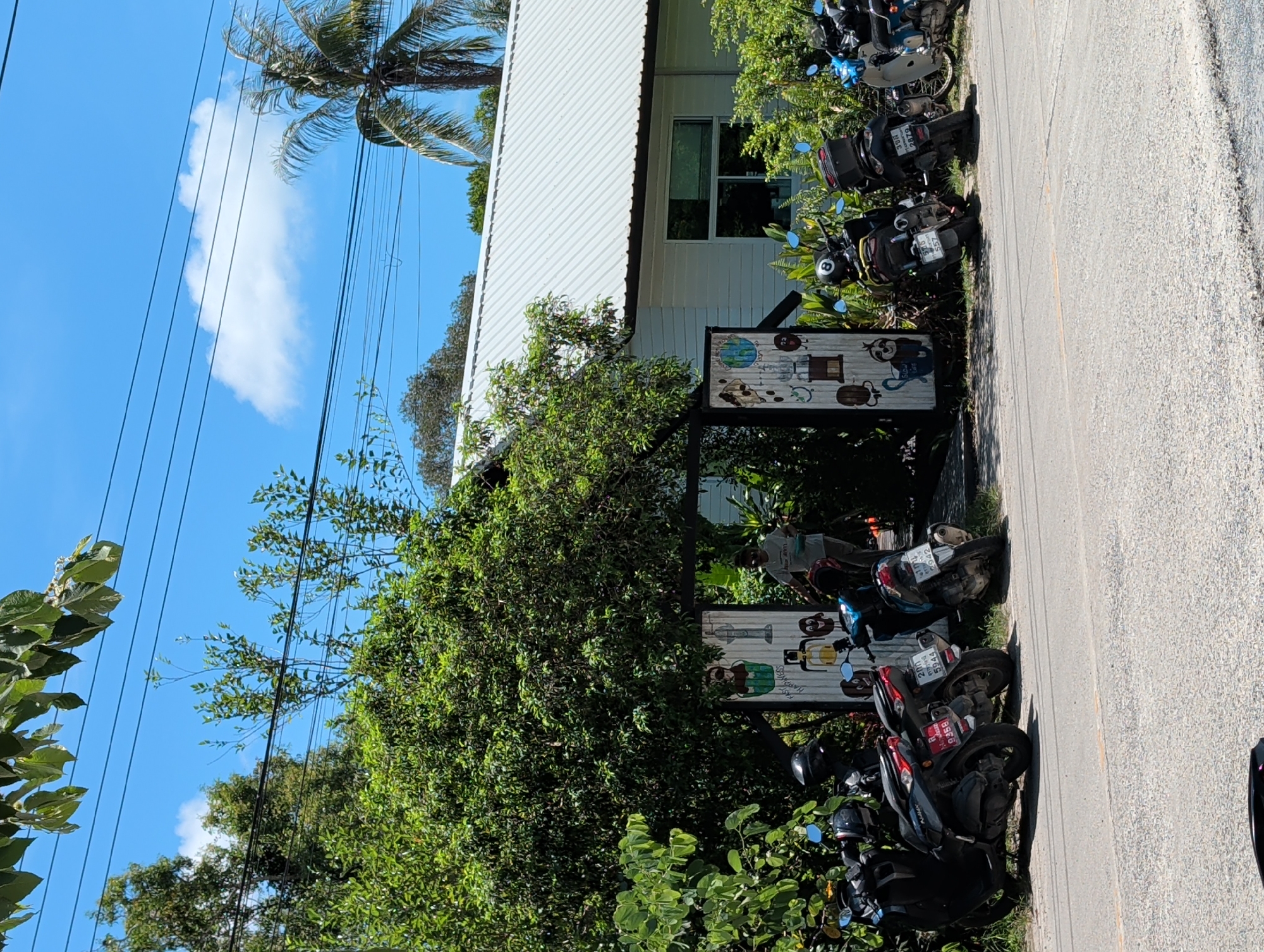 This photo was taken in ตำบลเกาะพะงัน, Thailand. It shows a building with a white exterior and a silver corrugated metal roof, partially obscured by trees. Two signs are visible in front of the building, each displaying cartoon illustrations. A row of parked motorbikes lines the edge of a road beside the building. The sky is blue with a few white clouds. Power lines are strung above. A palm tree can be seen to the left of the building. The road is light gray in color.
