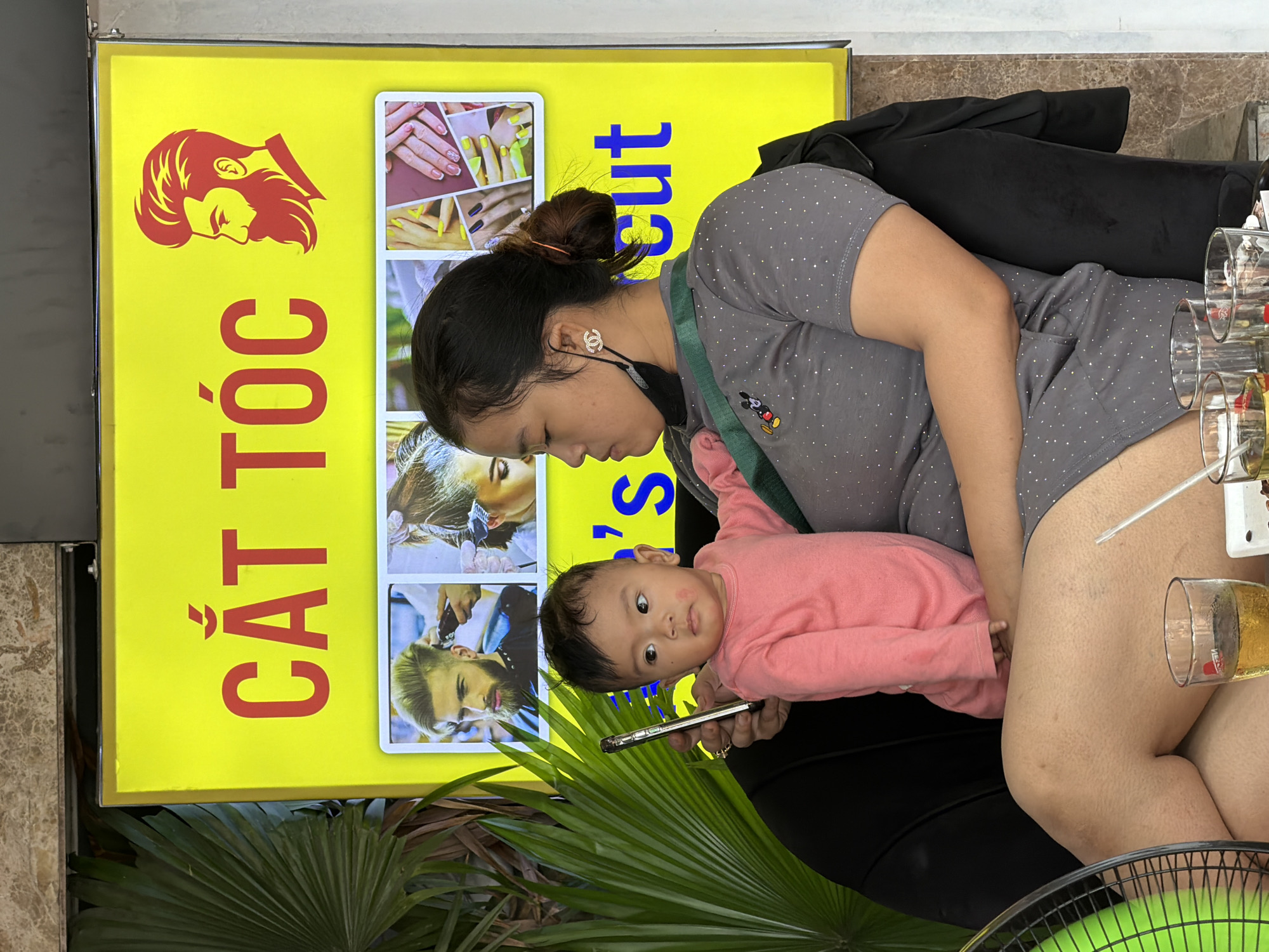 A bright daylight scene captures a woman and a baby seated outdoors in what appears to be a cafe or street-side establishment in Đà Nẵng, Vietnam. The woman, wearing a grey polka-dotted top and dark bottoms, holds a baby dressed in a pink long-sleeved shirt on her lap. She has dark hair tied in a bun, wears a Chanel-branded earring, and a Mickey Mouse pin on her shoulder strap. She is focused on a smartphone held in her hands. The baby, with dark hair and a round face, looks directly at the camera with wide, curious eyes.
Behind them is a prominent yellow signboard with red and blue text. Large red Vietnamese characters, oriented vertically, spell "CẮT TÓC," which translates to "HAIRCUT." Below this, "cut" is visible in blue letters, oriented horizontally. The signboard features images related to beauty services, including a stylized red silhouette of a man's head, pictures of manicured nails, and examples of hair styling and men's haircuts.
To the right of the woman, several glasses, some empty and one containing a light-colored beverage, are visible on a table. A clear straw protrudes from one. A black garment is draped over a chair or object next to the woman. In the foreground, a large green palm leaf and part of a green electric fan are visible, suggesting a warm climate. The ground or wall nearby is a light-colored marbled surface.