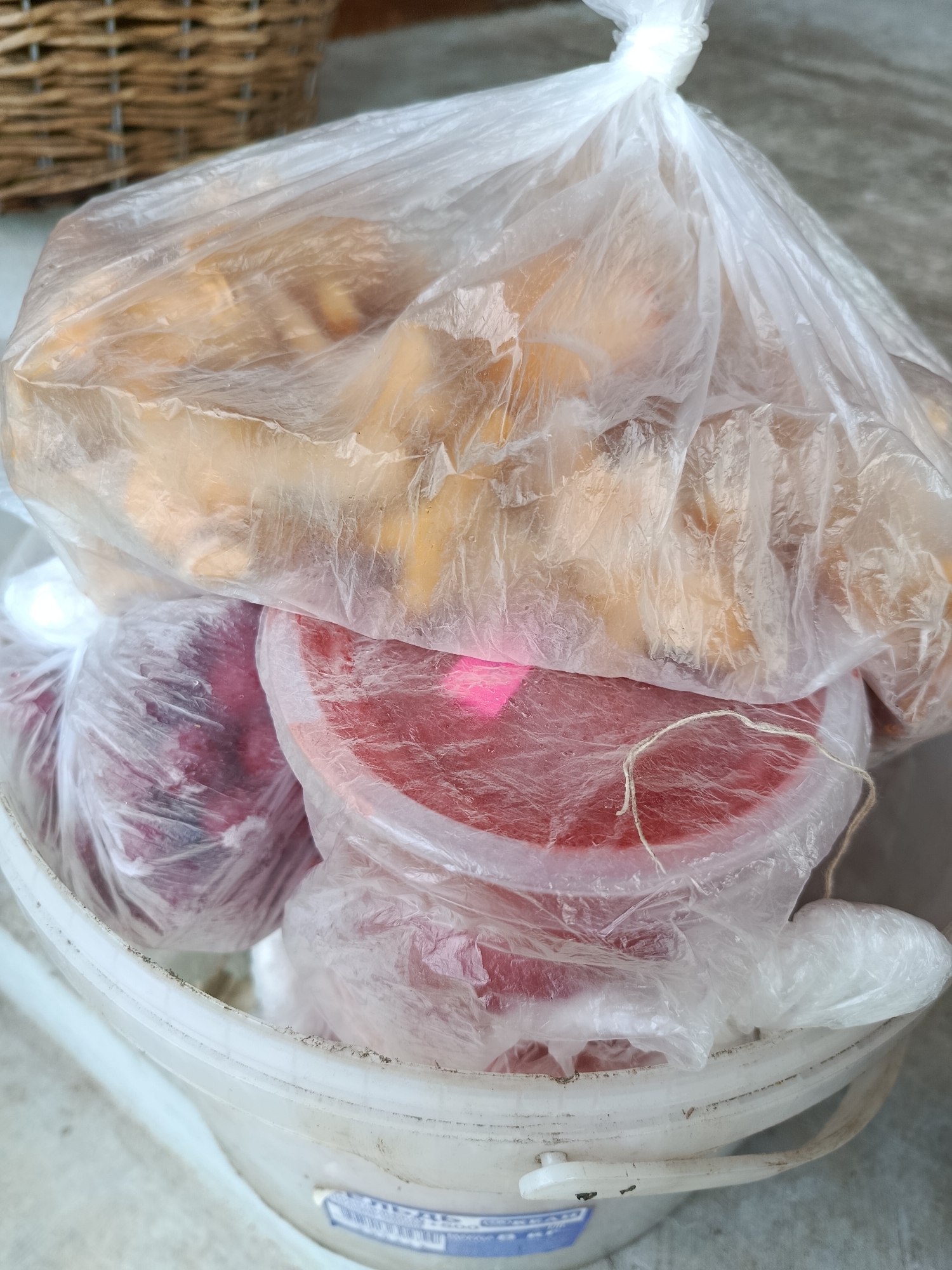 A bucket containing several plastic-wrapped items sits on a concrete surface. The bucket is white plastic with a blue and white label on the side. Inside the bucket, from bottom to top, are three plastic-wrapped items. The bottom item is a pinkish-red circular object, possibly a container with food, wrapped in clear plastic. Above this is a larger, round, red item, also wrapped in clear plastic and tied with string. The topmost item is a large, translucent plastic bag filled with what appears to be golden-brown, irregularly shaped food items, possibly fried dough or pastries. In the background, a portion of a wicker basket is visible. The overall scene suggests an outdoor or semi-outdoor setting, potentially a market or a storage area.