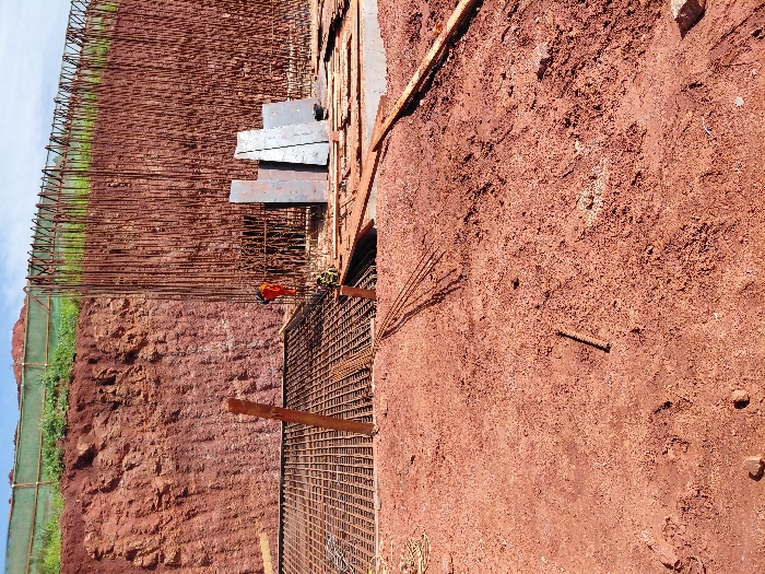 The image depicts an active construction site in Fdalate, Morocco, under a partially cloudy sky. The foreground consists of uneven, reddish-brown soil with scattered small debris and a horizontal steel rebar segment. To the left, a large, vertical cut of reddish-brown earth forms an excavated bank. Spanning the midground and right is a dense, vertical wall of steel rebar, with many bars bent into hooks at the top, indicating preparation for a concrete structure. At the base of this rebar wall, an orange object, possibly a piece of equipment, rests near a yellow object, potentially another piece of equipment or safety gear. Adjacent to this, to the left, is a flat area with a horizontal grid of rebar, likely for a foundation or slab. Further right, a stack of four dark, rectangular metal sheets is positioned on a low concrete base. Wooden planks are scattered on the ground and embedded near the rebar structures. The top of the excavated bank in the background is covered with green vegetation secured by a light green mesh fence. No individuals are present in the scene.