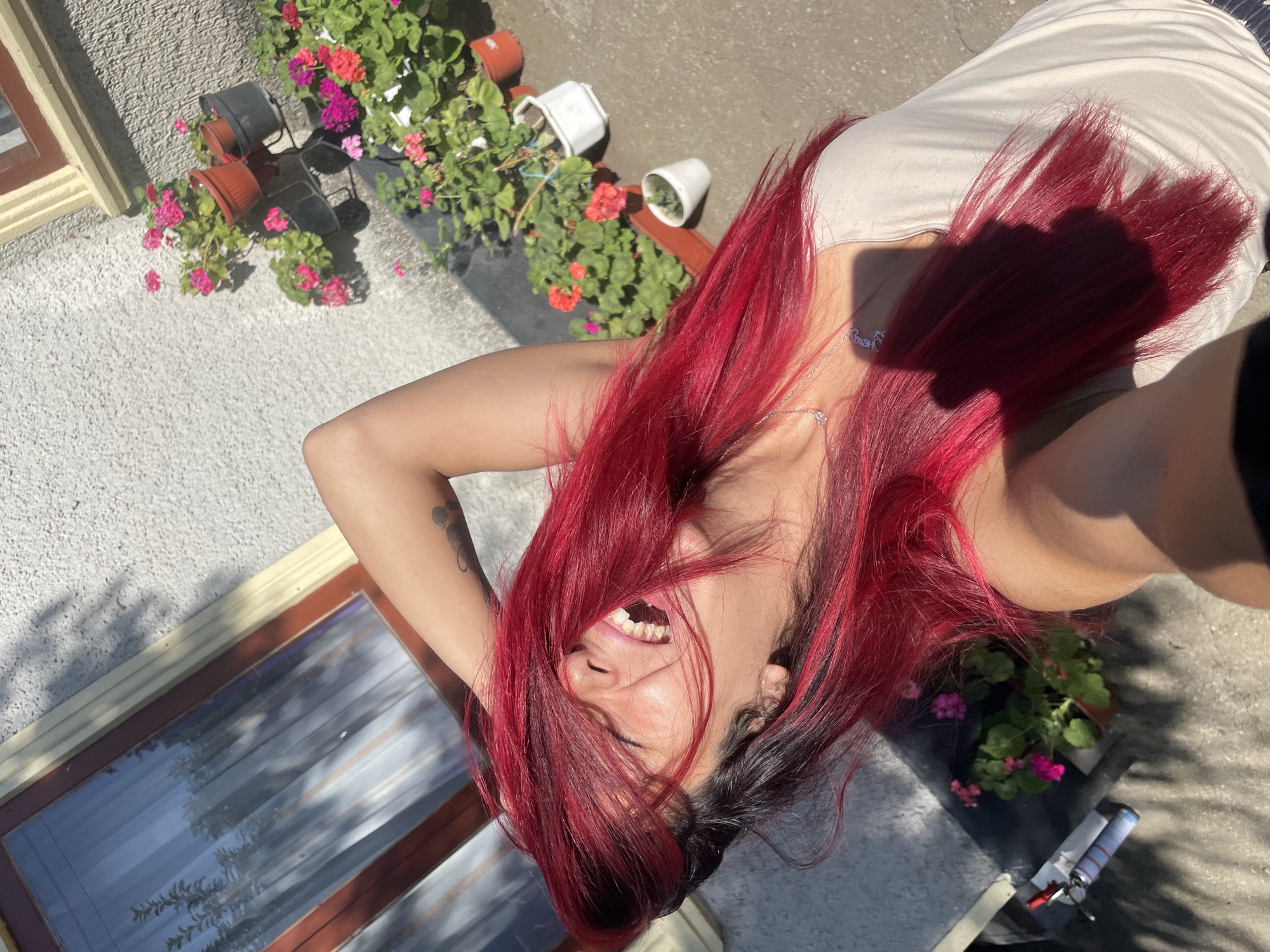 In Zheglartsi, Bulgaria, a young woman with vibrant red hair laughs with her head tilted back, her right arm raised and hand in her hair. She is outdoors, in what appears to be a residential courtyard, with a textured, light-colored wall and a window with sheer curtains visible behind her. Sunlight casts shadows across the scene, highlighting a collection of potted flowers, including red and pink geraniums, arranged on tiered stands against the wall. A concrete patio or ground is visible in the foreground, with a shadow of the photographer falling across the woman's shoulder and chest. The woman is wearing a light-colored tank top and a silver necklace.