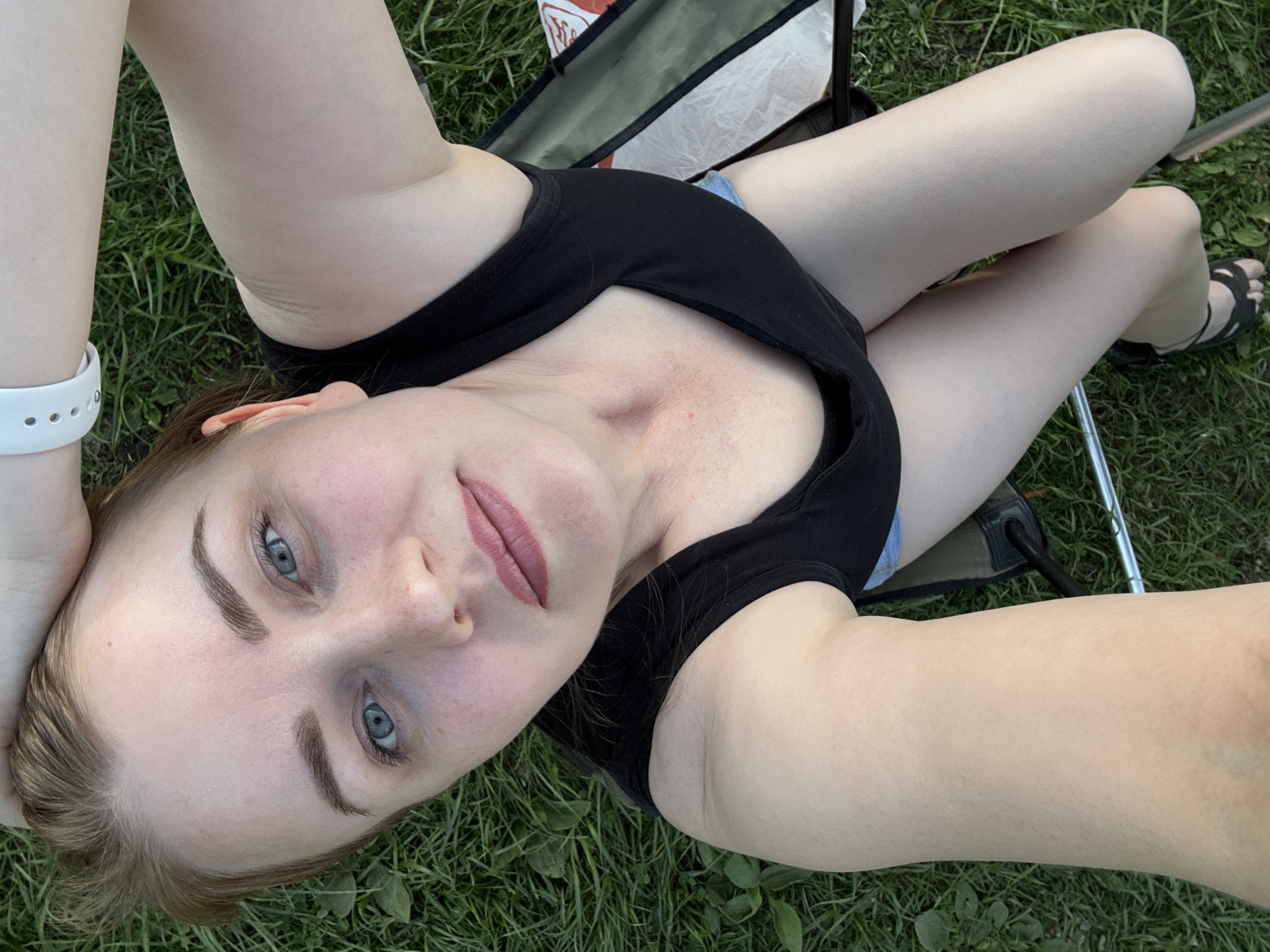 A woman with light skin, light brown hair, and blue eyes is lying on her back in a grassy area. She is wearing a black tank top and what appears to be blue shorts. Her legs are crossed. She is wearing a white wristband on her left wrist and black sandals on her feet. A camping chair is partially visible behind her. The location is presumed to be Chaikovsky, Russia, based on the prompt.