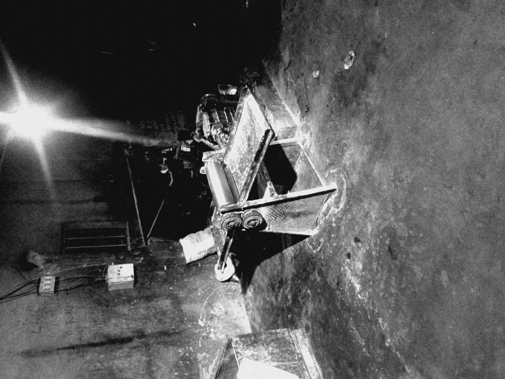 The image depicts a dimly lit, confined space in an unknown location. A metal cart with multiple levels and a cylindrical object rests on the right side of the frame, casting a shadow on the textured wall. To the left, there is a light source, possibly a flashlight, illuminating the scene. In the background, various objects, including boxes, electrical outlets, and what appears to be graffiti on the wall, can be observed. The floor is rough and uneven. No individuals are directly visible in the image.