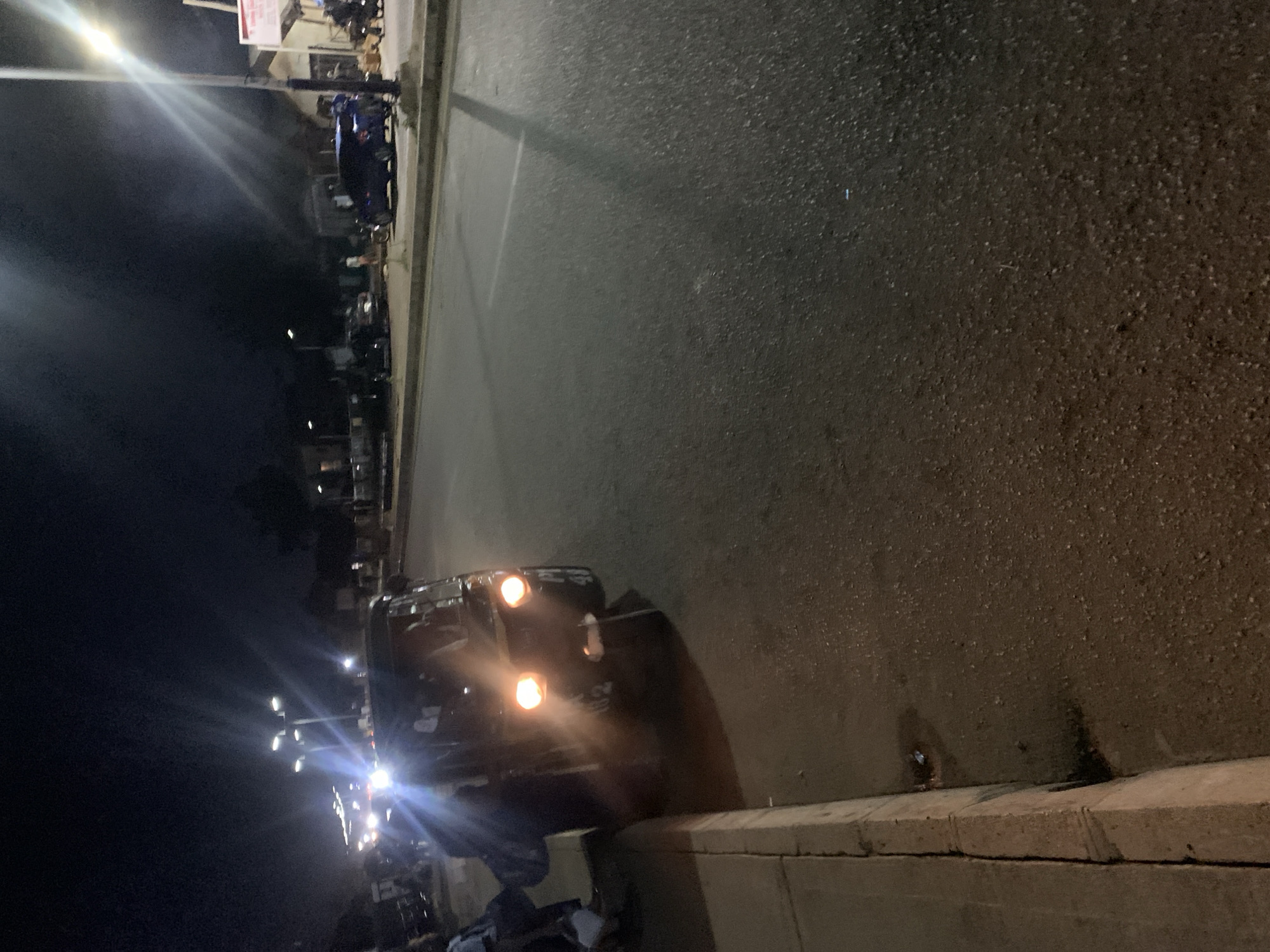 The image depicts a nighttime scene at an unidentified location in New Stadium, Nigeria. A dark-colored vehicle is parked on the roadside, illuminated by its headlights. In the background, a row of streetlights lines the road, casting a bright glow over the area. Additional vehicles and individuals are visible further down the street, suggesting a bustling urban environment. The foreground shows a concrete curb bordering the road.