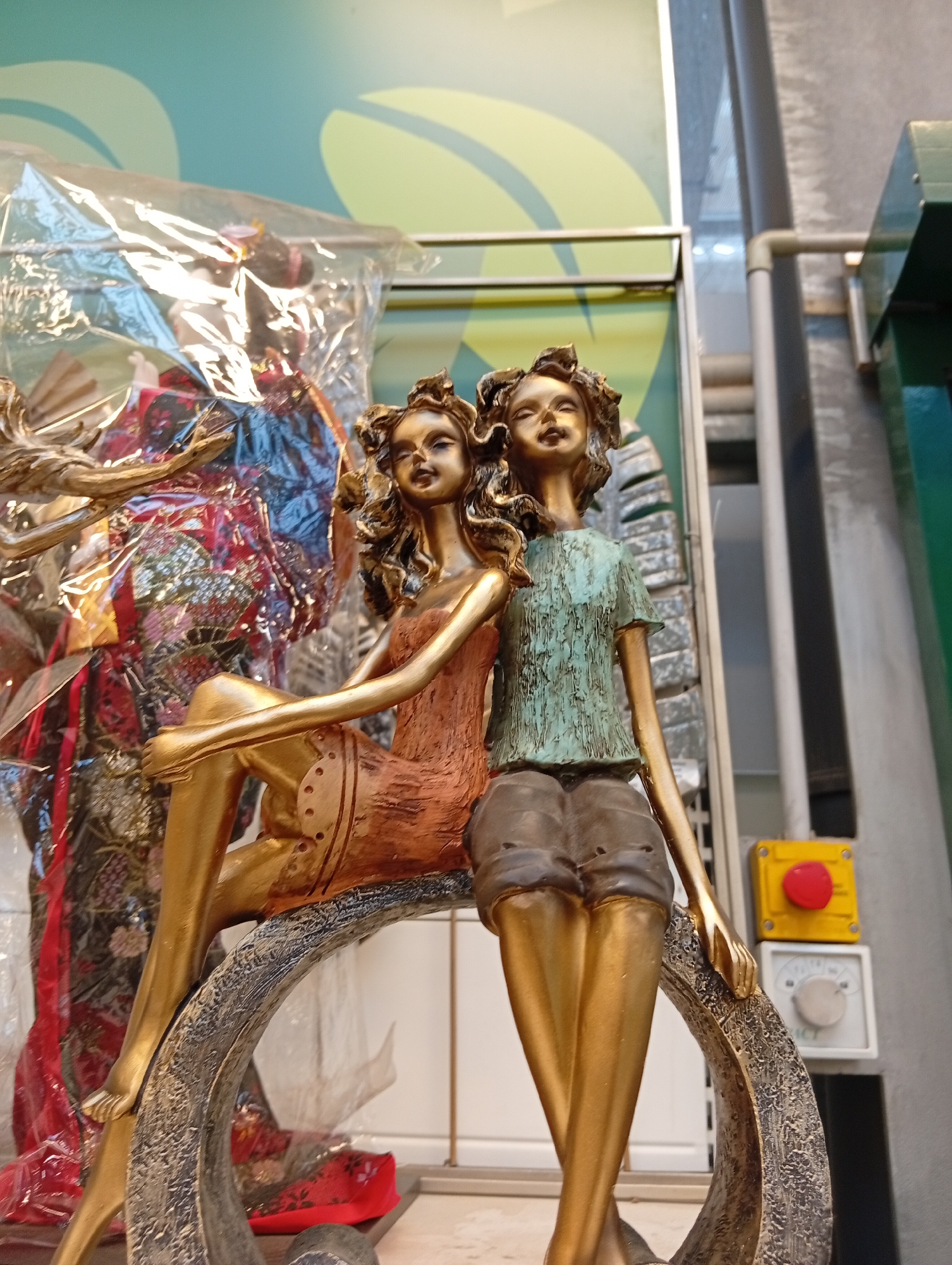 A golden sculpture depicts two female figures sitting together on a curved object. The figure on the left wears an orange-brown textured dress, and the figure on the right wears a light blue, textured t-shirt and brown shorts. Both figures have curly hair and closed eyes, suggesting relaxation or contemplation. In the background, partially obscured by plastic, is a colorful display, possibly of traditional Japanese dolls or fabric. A yellow electrical box with a red button is visible on the right side of the image, mounted on a wall. The scene appears to be indoors, potentially in a store or exhibition space. The setting does not provide specific clues to its location in Pushkino, Russia.