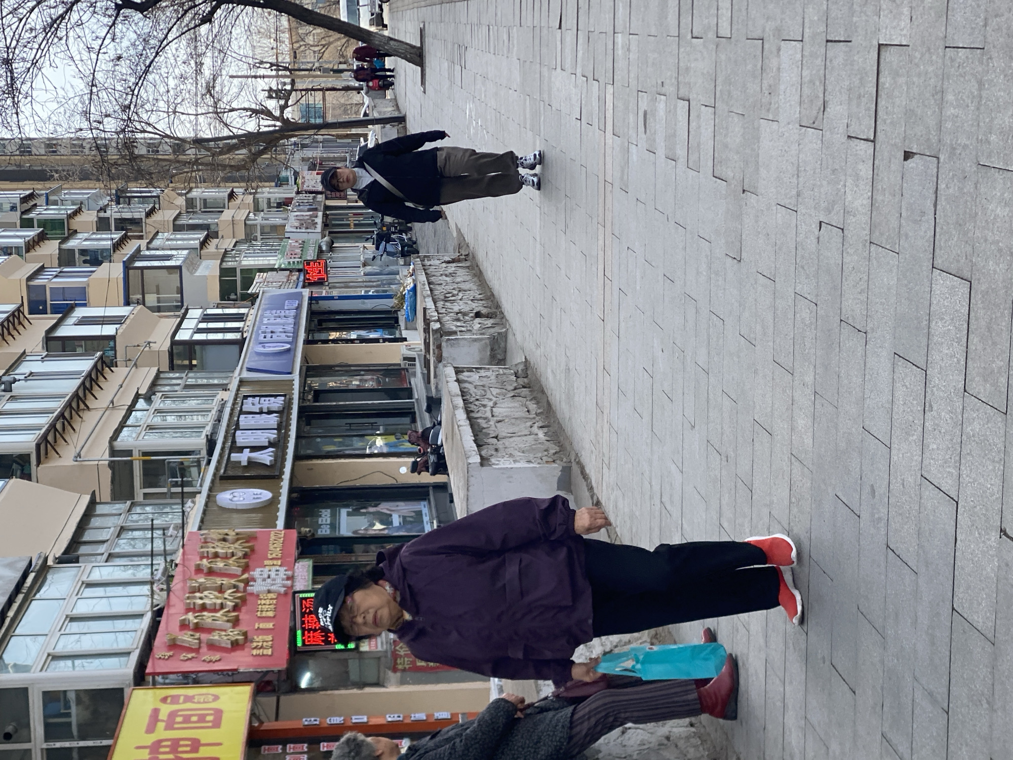 The image shows a street scene in what appears to be Zhongtun, China. Along the left side of the image are buildings with many windows, along with shops and restaurants with their signs visible. Pedestrians can be seen on the sidewalk. In the foreground, two older women are walking. One is wearing a purple jacket and black pants with red sneakers. The other woman is in a gray coat with striped pants and red shoes. Another person is walking on the other side of the sidewalk, wearing a black jacket and khaki pants. The sidewalk is made of light gray paving stones.