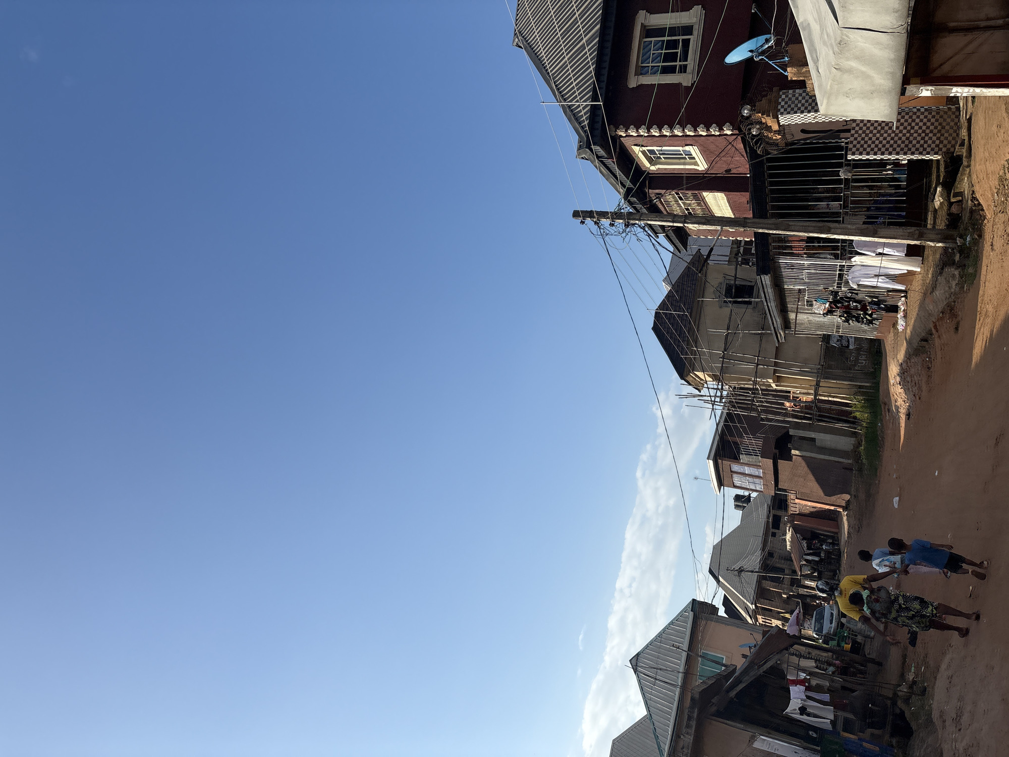 The image depicts a residential street scene in Obosi, Nigeria, under a clear blue sky. In the foreground, a woman with a child on her back and two other children walk down a dirt road. They are heading away from the camera, towards a cluster of buildings.
The buildings lining the street are a mix of multi-story structures, some appearing to be under construction or in disrepair, with corrugated metal roofs and exposed scaffolding. One prominent building on the right is painted a deep red with white decorative trim around its windows. A satellite dish is mounted on its exterior. In front of this building, white garments are hanging on display behind a metal gate.
The street itself is unpaved and appears dusty. Utility poles with a network of wires are visible along the street, connecting the buildings. The overall impression is one of a densely populated urban environment with evidence of ongoing development and daily life.