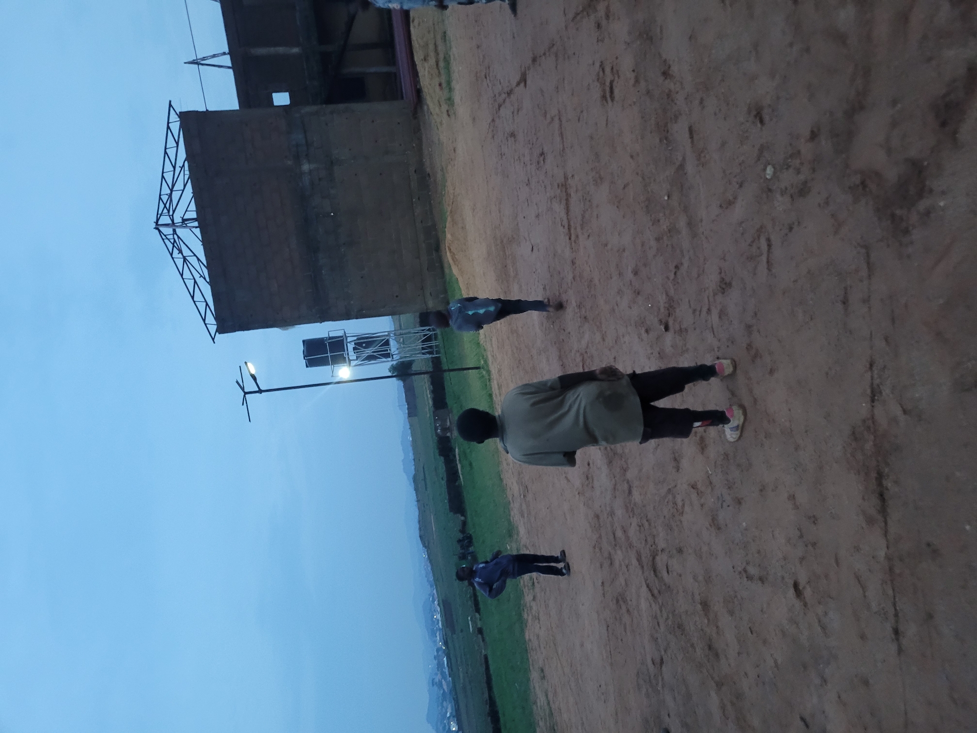 An outdoor scene captures three young males in the foreground and midground on a dirt surface, with a distant hilly landscape. The sky is a deep twilight blue, with a single street lamp visible in the upper left quadrant. To the right, a partially constructed brick building with a metal roof frame stands against the sky. In the middle ground, a young male in a grey t-shirt and black shorts is facing away from the camera. To his left, another young male in a blue shirt and dark pants stands with hands on his hips. Further to the left, a third young male in a dark shirt and dark pants is also visible. A water tower is present behind the partially constructed building. The ground is composed of loose dirt with some vegetation in the midground.