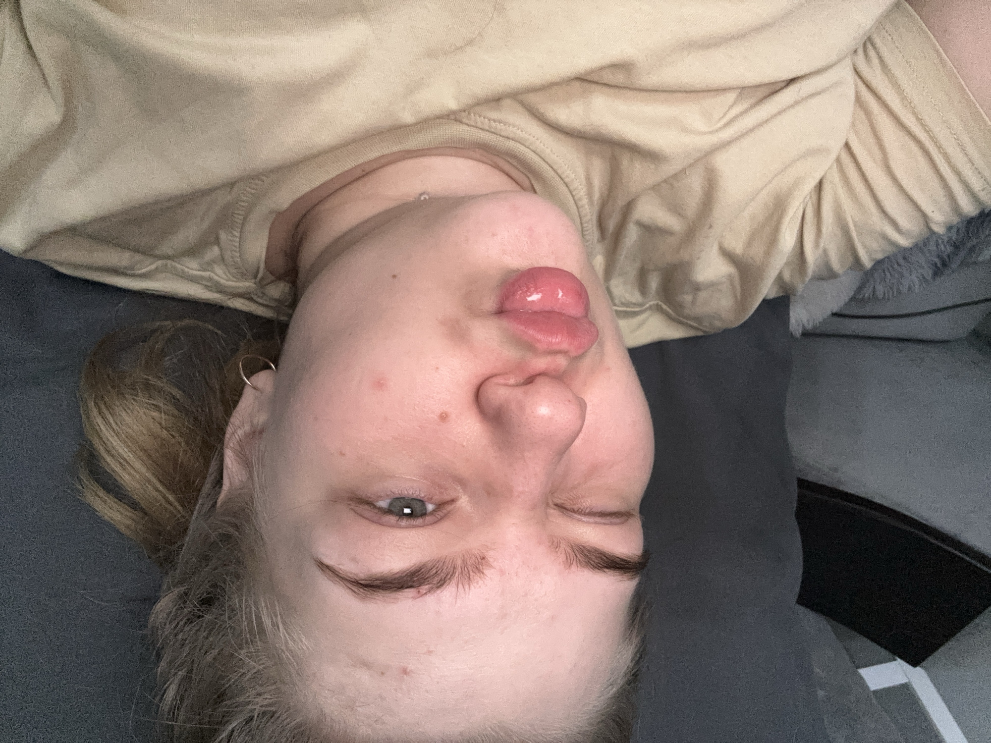 A close-up, upside-down selfie of a fair-skinned person with light brown hair. The person is sticking their tongue out and winking their right eye. They are wearing a tan-colored shirt or top. The background appears to be a gray surface, likely a couch or bed. There is no verifiable information to place this scene in Chaikovsky, Russia.