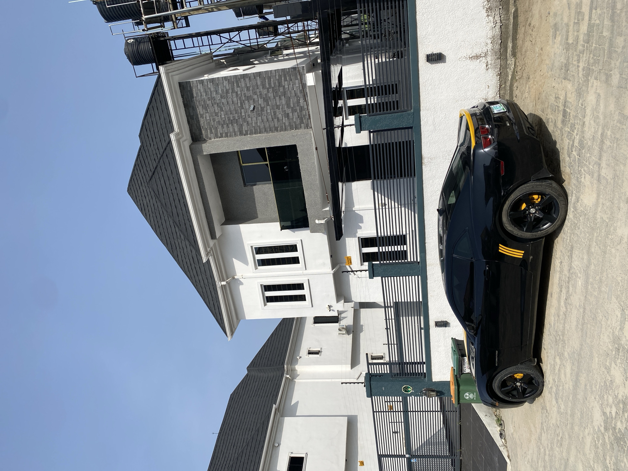 The image presents an exterior view of a modern residential property in Lagos, Nigeria, characterized by a two-story white building with a gray roof. The property is enclosed by a black metal fence. A black sports car with yellow stripes is parked in front of the building. Overhead water tanks on a metal structure are visible above the building. The environment suggests a clear sky and a sunny day. A green and yellow trash can sits at the curb near the fence. There are no people visible in the image.