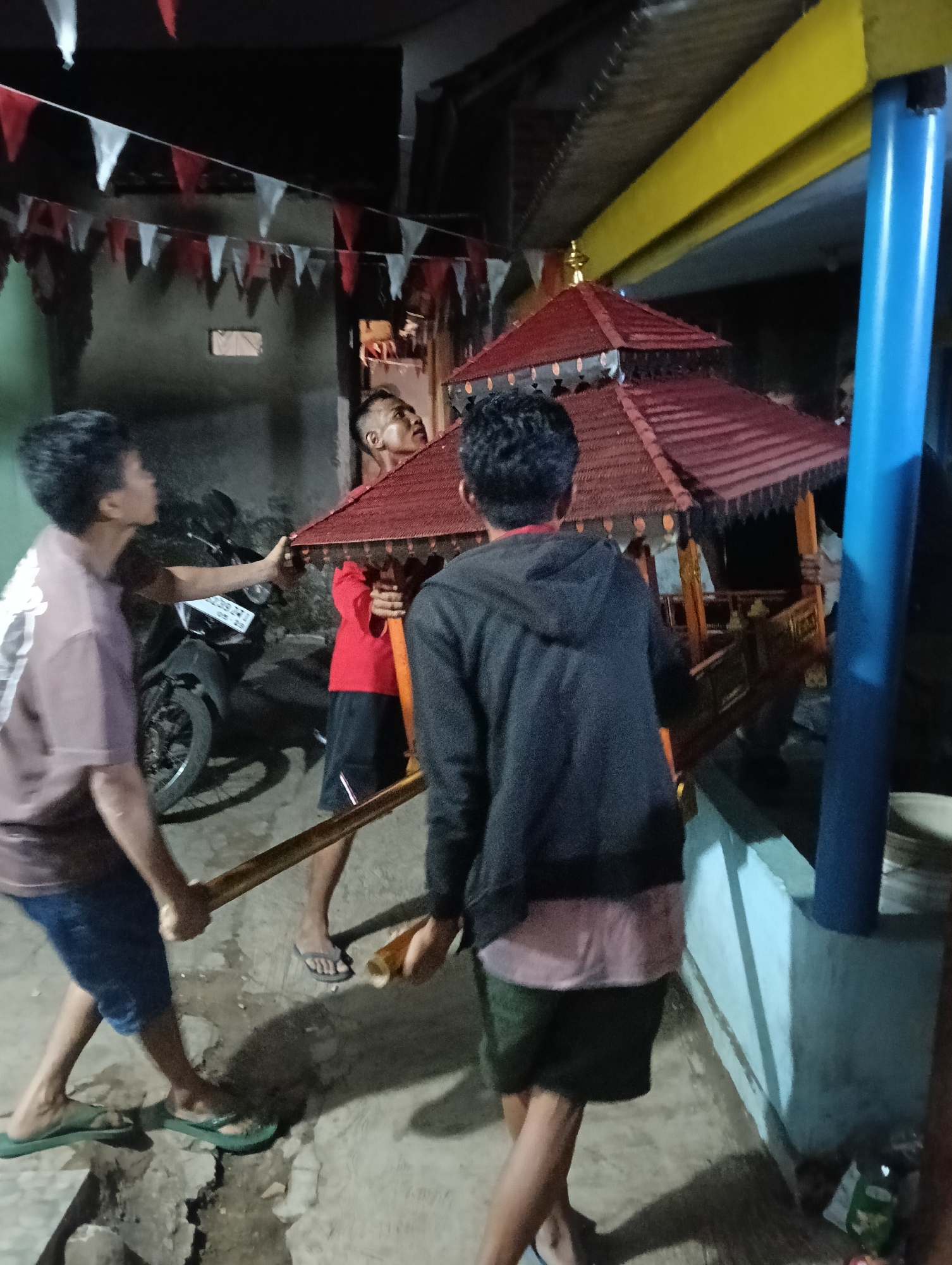 In the city of Tasikmalaya, Indonesia, three young men are carrying a small, ornate, red-roofed structure, possibly a replica of a temple or pavilion, on wooden poles. The structure has intricate carvings and a golden finial. They are walking on a dirt and concrete ground, with some cracks visible. The scene appears to be outdoors at night, with overhead red and white pennants strung across the area, suggesting a festive occasion or cultural event. A motorcycle is parked in the background. One of the men is wearing a purple t-shirt and blue shorts, another a red t-shirt and black shorts, and the third a black hooded sweatshirt and green shorts. The lighting is dim, with artificial light sources casting shadows.
