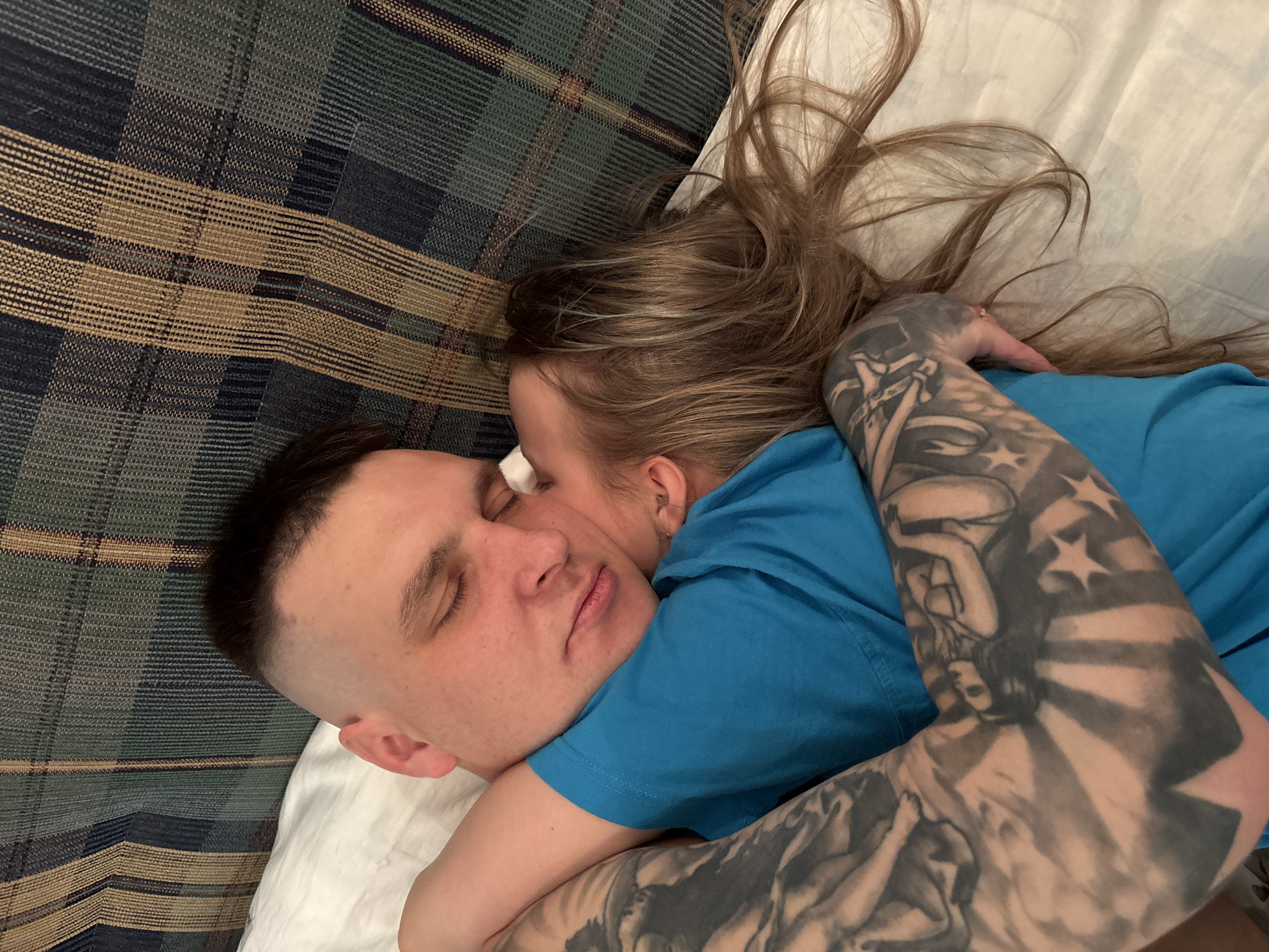 An image depicts two individuals lying embraced on a bed. An adult male, positioned on his back, has short dark hair with a faded buzz cut on the sides, and his eyes are closed. He is wearing a blue t-shirt. His right arm is visible, covered in extensive black and grey tattoos depicting figures and stars. An adult female is curled against his right side, her face pressed against his cheek, eyes also closed. She has long, light brown hair spread across the white bedding and a blue t-shirt. Her right arm is wrapped around the male's torso. The bed features white sheets and pillows. Behind the subjects, a patterned textile, possibly a blanket or headboard, exhibits a plaid design with vertical and horizontal stripes in dark blue, green, and beige tones. The scene is set indoors, within what appears to be a bedroom, assumed to be in the city of Русский Вожой, Russia.