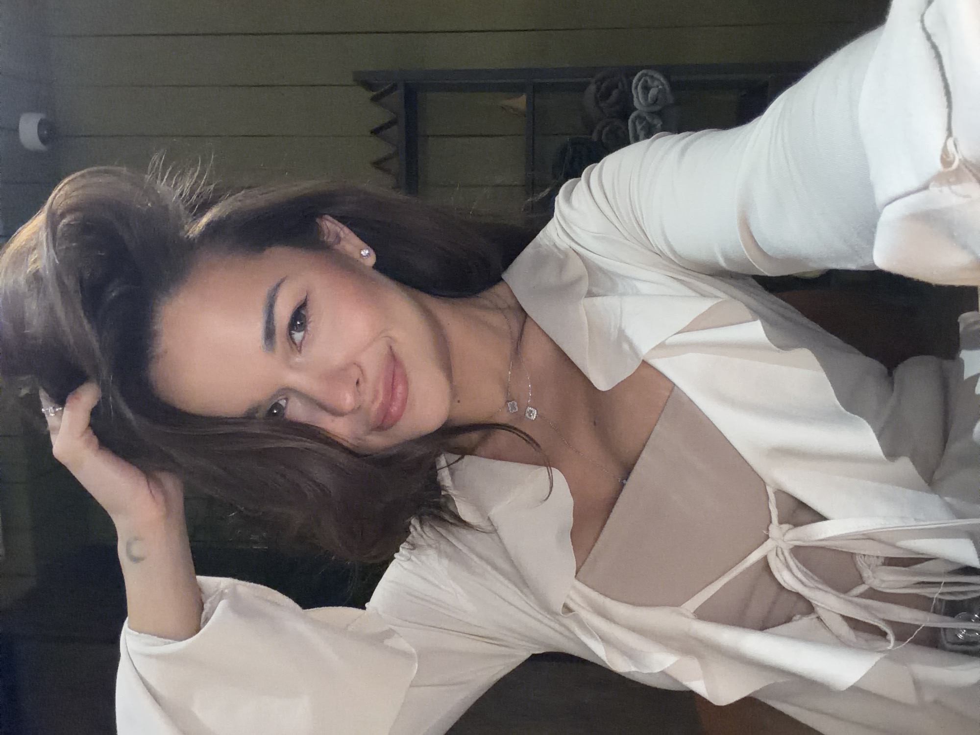 The image is a selfie of a woman indoors. She has brown hair and fair skin and is wearing a white top and a necklace. She is holding her head in her hand and smiling slightly. In the background, there is a shelf with rolled-up towels and a security camera. The location is unknown.