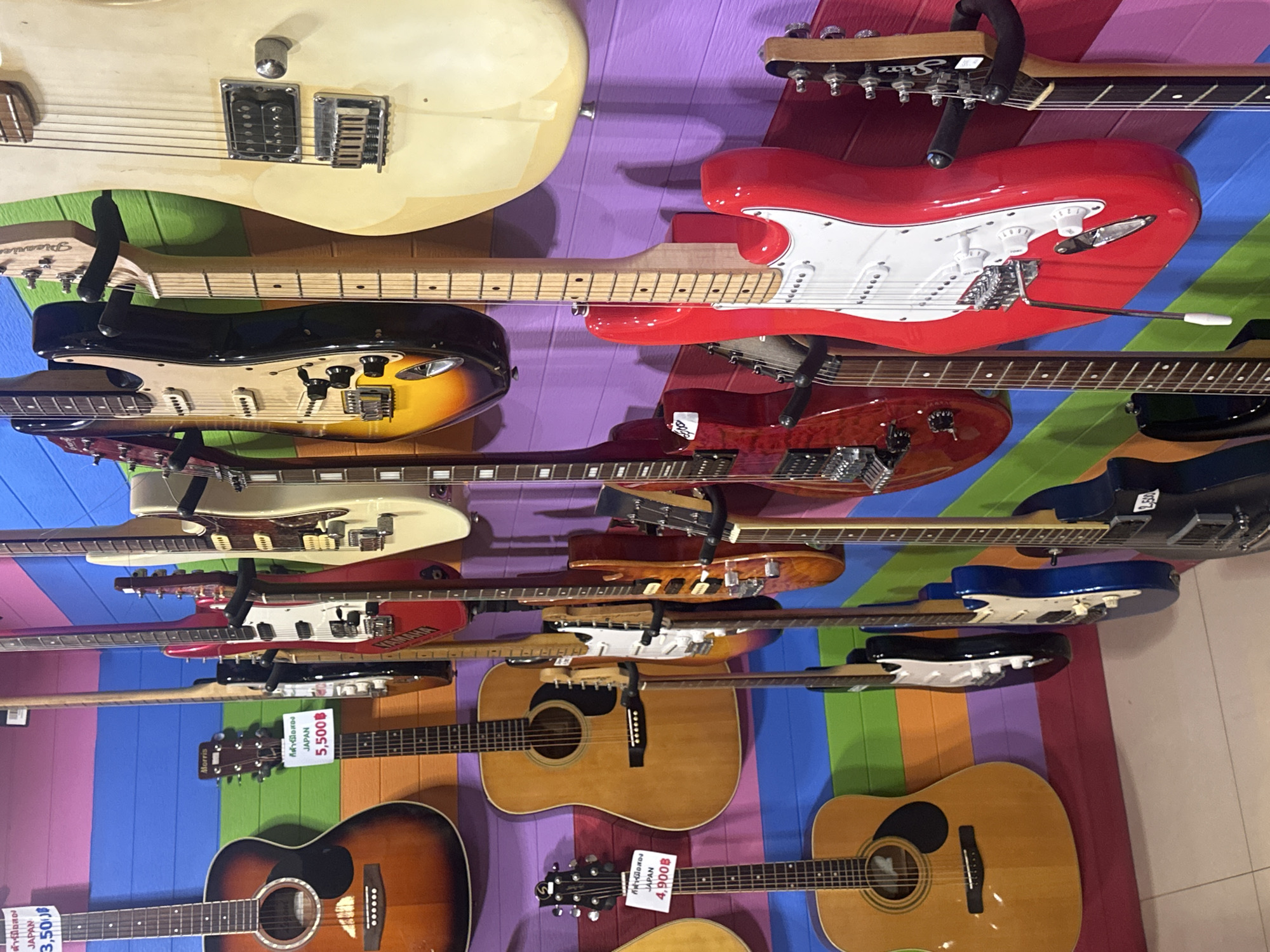 The image depicts a vertical display of electric and acoustic guitars mounted on a wall, likely in a music store. The wall is painted with alternating vertical stripes of purple, green, blue, and pink, creating a colorful backdrop for the guitars. The guitars are arranged in a stacked manner, with various models and colors visible, including red, cream, black, sunburst, and natural wood finishes. Price tags are affixed to some of the guitars, indicating that they are for sale. The tags contain the word "JAPAN" and a price in Thai Baht, suggesting the instruments might be used guitars from Japan. Additional brand names such as "Sire" and "Picaric" are visible. The environment suggests a retail setting for musical instruments, possibly in Pattaya City, Thailand.