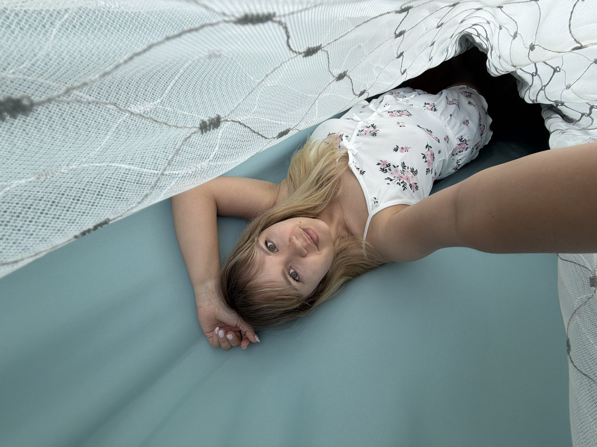 A young fair-skinned woman with blonde hair and blue-green eyes lies on a blue-green surface, possibly a bed or a large pad. She wears a white floral-patterned top. She's taking a selfie with her right arm outstretched. The scene is partially obscured by a white, lacy curtain, creating an enclosed or tent-like feeling. The photo was likely taken in a residential interior in Chaikovsky, Russia.
