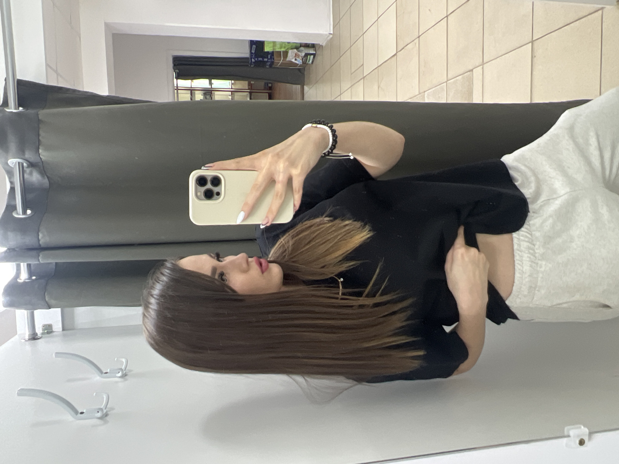 A young woman with long, straight brown hair, bangs, and dark eyeliner is taking a mirror selfie in what appears to be a changing room or a small retail space. She is wearing a black t-shirt and light gray sweatpants. She is holding a light-colored smartphone in her right hand, and her left hand is touching her stomach area. She is wearing a bracelet on her right wrist. The background consists of a plain white wall with two white coat hooks, a dark gray curtain, and a tiled floor. In the reflection of the mirror, a shelf with items and another curtain can be seen. The overall lighting is bright. There is no visual information to suggest this image was taken in Noviy, Russia.
