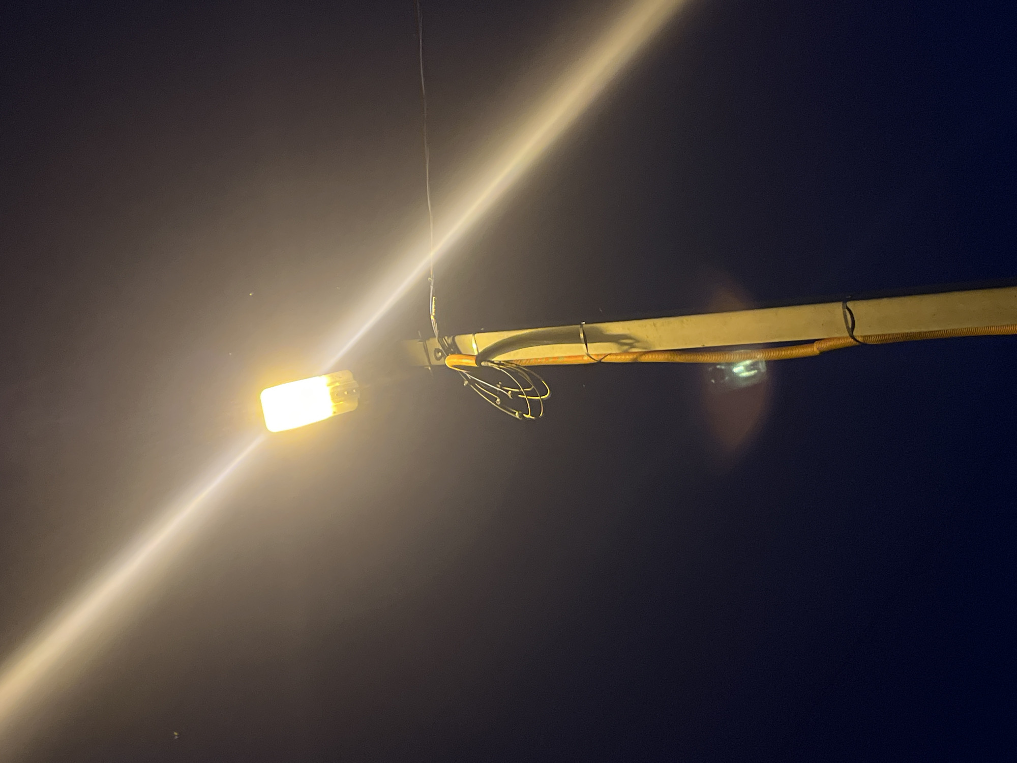 The image depicts a street lamp at night in the city of Nizhny Tagil, Russia. The lamp is emitting a bright yellow light that beams diagonally across the dark sky. A portion of the lamp post is visible, made of concrete with a yellow conduit running along its side. Some electrical wires are attached to the post, with a coiled wire harness visible near the top. The background is a dark, hazy night sky, with some subtle light pollution creating a gradient of darkness. There are no people or other discernible subjects in the frame. The overall scene is atmospheric and captures the solitary illumination of a street light against the night.