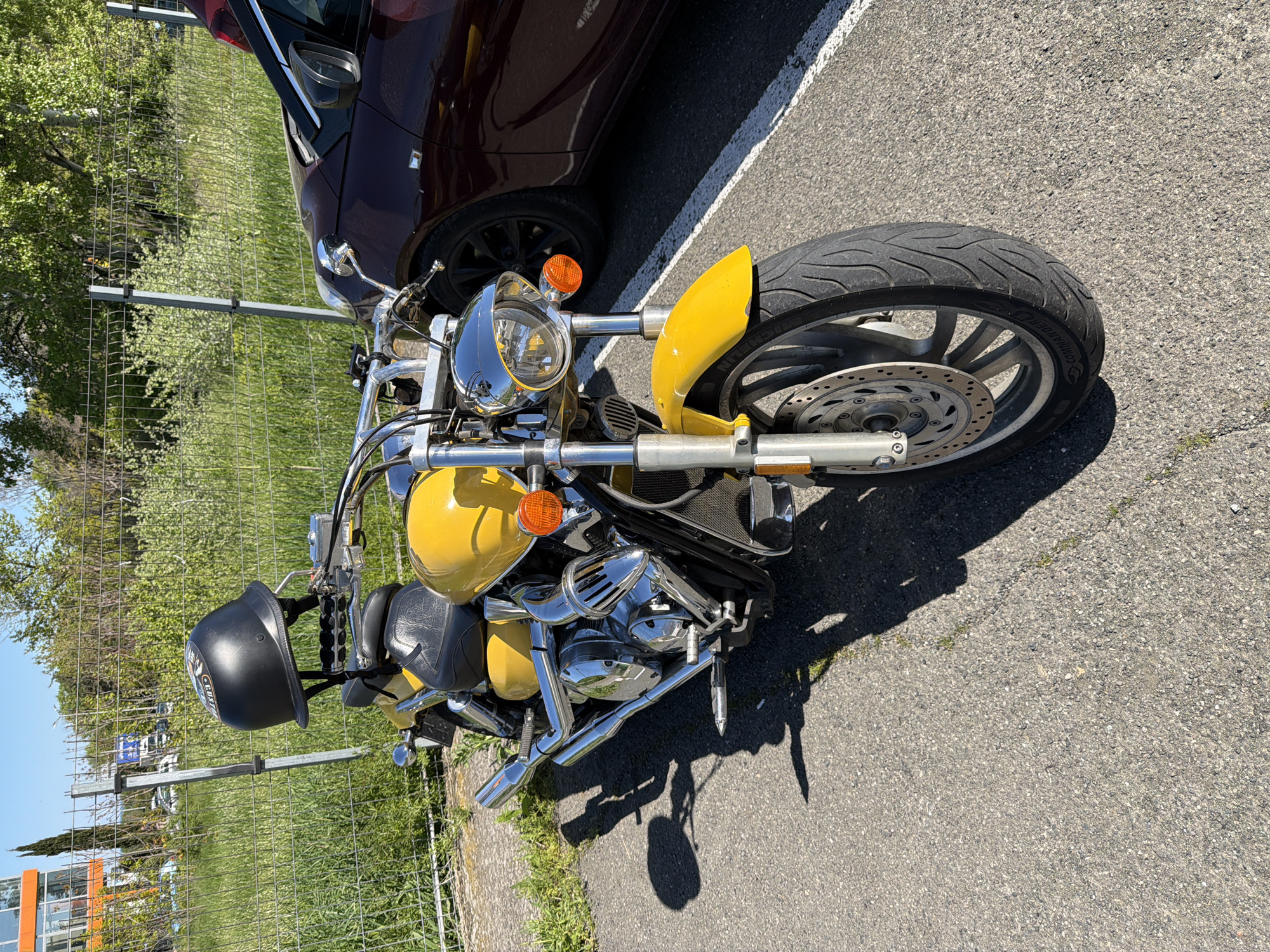 A yellow motorcycle is parked on an asphalt surface next to a maroon car. The motorcycle has chrome accents and a black helmet is attached to the rear. A metal fence and green vegetation are visible in the background. The scene is outdoors on a sunny day.