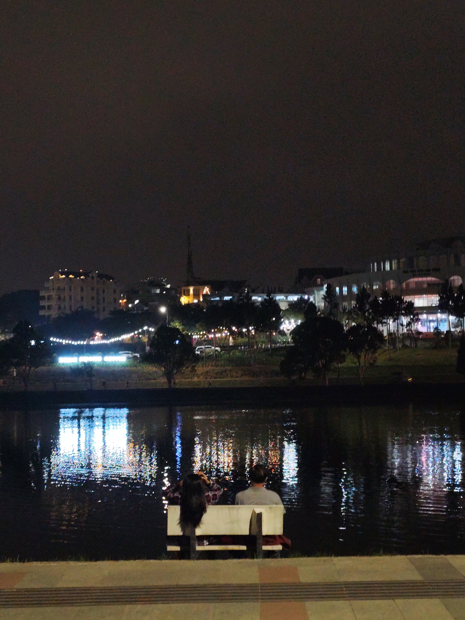 The image shows a nighttime view of Da Lat, Vietnam, featuring a body of water reflecting city lights. In the foreground, a couple sits on a bench facing the water. The city skyline includes buildings and a steeple illuminated by lights, with decorative lights strung between trees. The water surface is speckled with reflections from the city lights. The foreground pavement is tiled.
