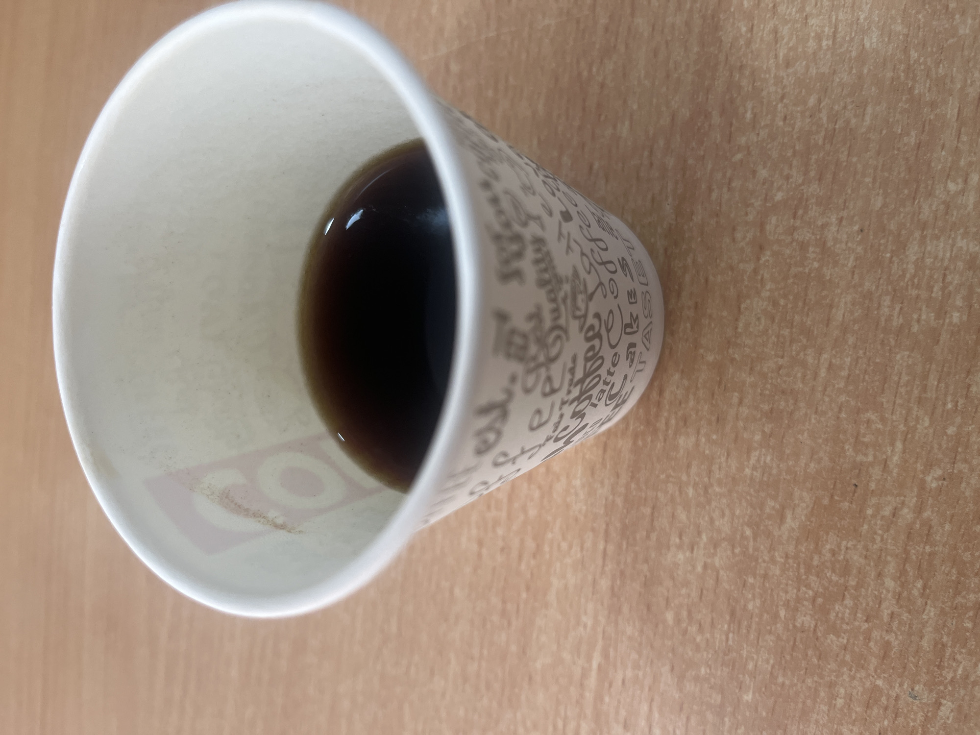 The image shows a partially filled white paper cup on a light brown, textured surface, possibly a desk or table. The cup contains a dark liquid, likely coffee or tea. The cup is decorated with a pattern of text and small symbols. The scene appears to be indoors. There is no evidence to indicate the location is Yeghegnadzor, Armenia. No people, actions, or interactions are visible.