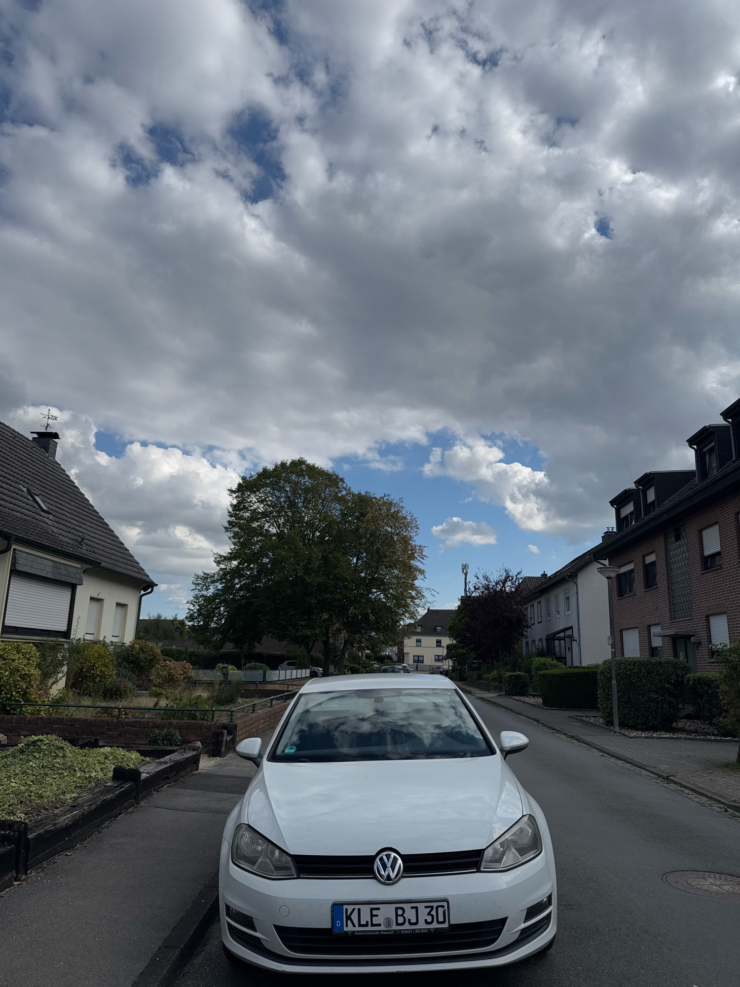 A white Volkswagen Golf is parked on the right side of a street in Cleves, Germany. The car is facing forward, and its license plate reads "KLE BJ 30". The street is lined with houses on both sides, and there is a large tree to the left of the car. The sky is cloudy, with patches of blue visible. The overall scene depicts a residential street on a cloudy day.