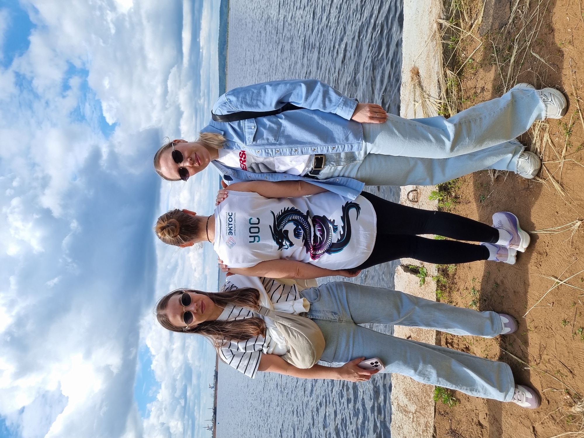 Three young women stand outdoors by a body of water under a cloudy sky. The woman on the left wears a striped t-shirt, light blue jeans, and light-colored sneakers. She has long brown hair and wears oval sunglasses. A beige crossbody bag is slung over her shoulder, and she holds a pink smartphone. The woman in the center has her back to the camera and wears a white sleeveless top with a dragon graphic and Russian text, black leggings, and purple sneakers. Her hair is pulled up in a bun. The woman on the right wears a light blue denim shirt over a white t-shirt with red text, light blue jeans, a black belt, and white sneakers. She has blonde hair pulled back and wears oval sunglasses. The group is positioned on a sandy bank next to a concrete ledge, with a wide river or lake behind them. In the distance, industrial structures and ships are visible on the horizon. The overall scene suggests a casual outing near a waterfront in the city of Chaikovsky, Russia.