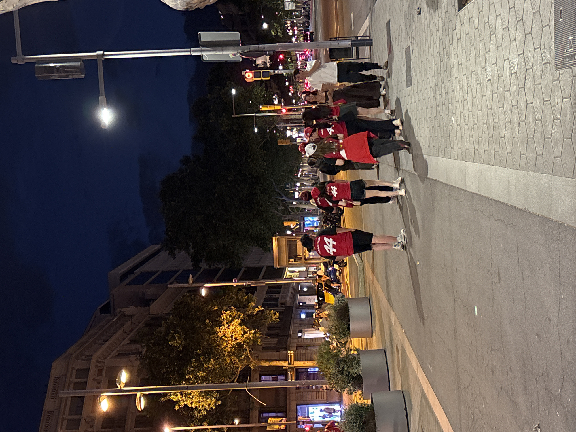 The image depicts a street scene at night in Barcelona, Spain, likely on a pedestrian walkway adjacent to a road. A group of people, predominantly young adults, are walking along the sidewalk. Several individuals are wearing matching red shirts with the number "44" and the name "HAMILTON" visible on the back, suggesting they are part of a fan group or attending an event. One person in the foreground wears a red shirt with "44" and black shorts, while another in the background wears a similar shirt with white shorts. A man in a white t-shirt and dark pants is seen walking away from the group.
The environment features streetlights illuminating the walkway and the adjacent road, where traffic, including scooters and cars, is visible. Buildings line the street, with some showing illuminated windows. Planters with greenery are placed along the sidewalk. The overall atmosphere is casual and suggests an urban setting during the evening.