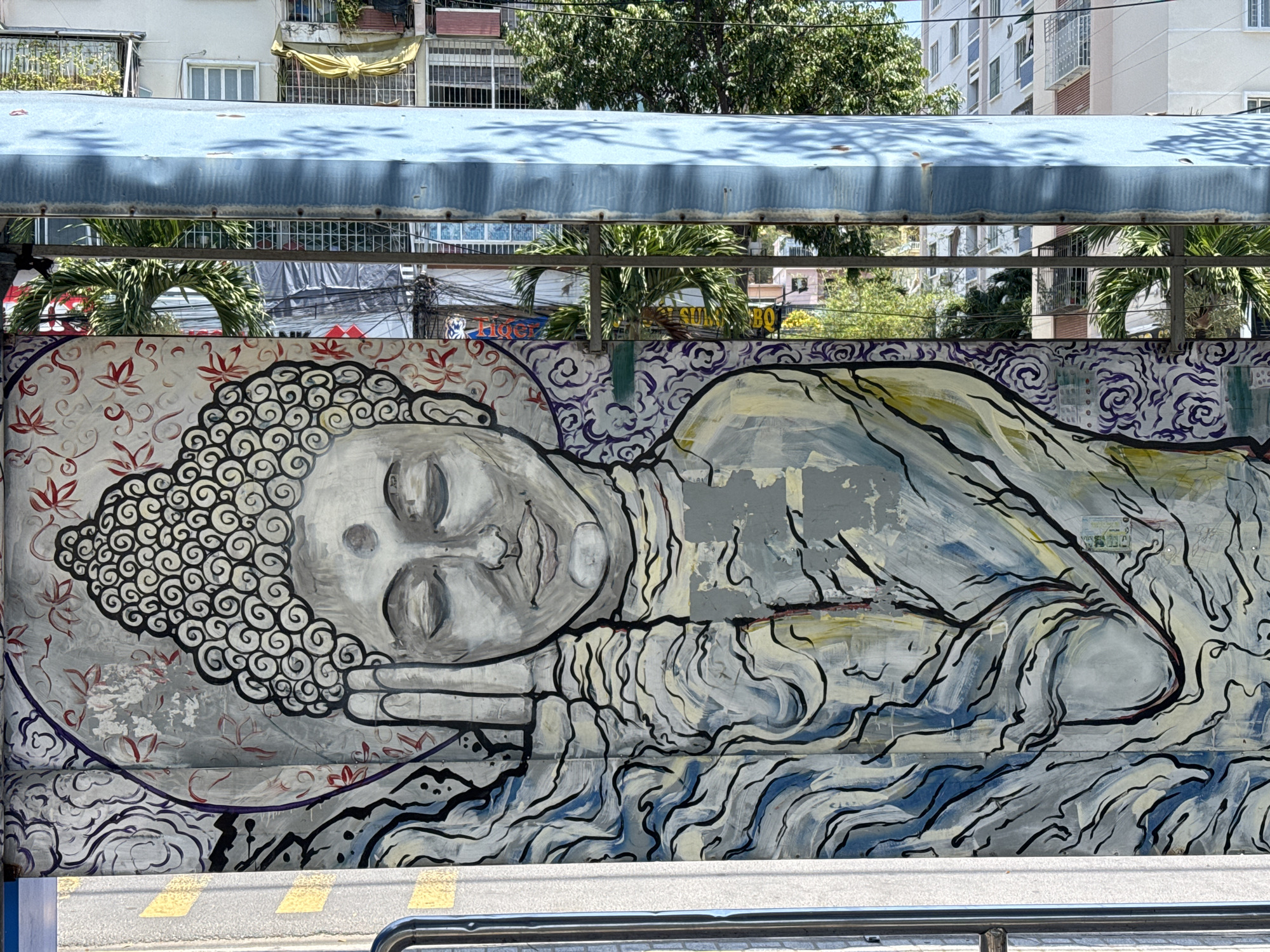 This photograph shows a mural of a reclining Buddha. The mural is painted on a wall, which is visible behind a bus shelter in Bac Nha Trang Ward, Vietnam. The background includes buildings and trees. The mural is rendered in gray, black, and white tones with some blue.