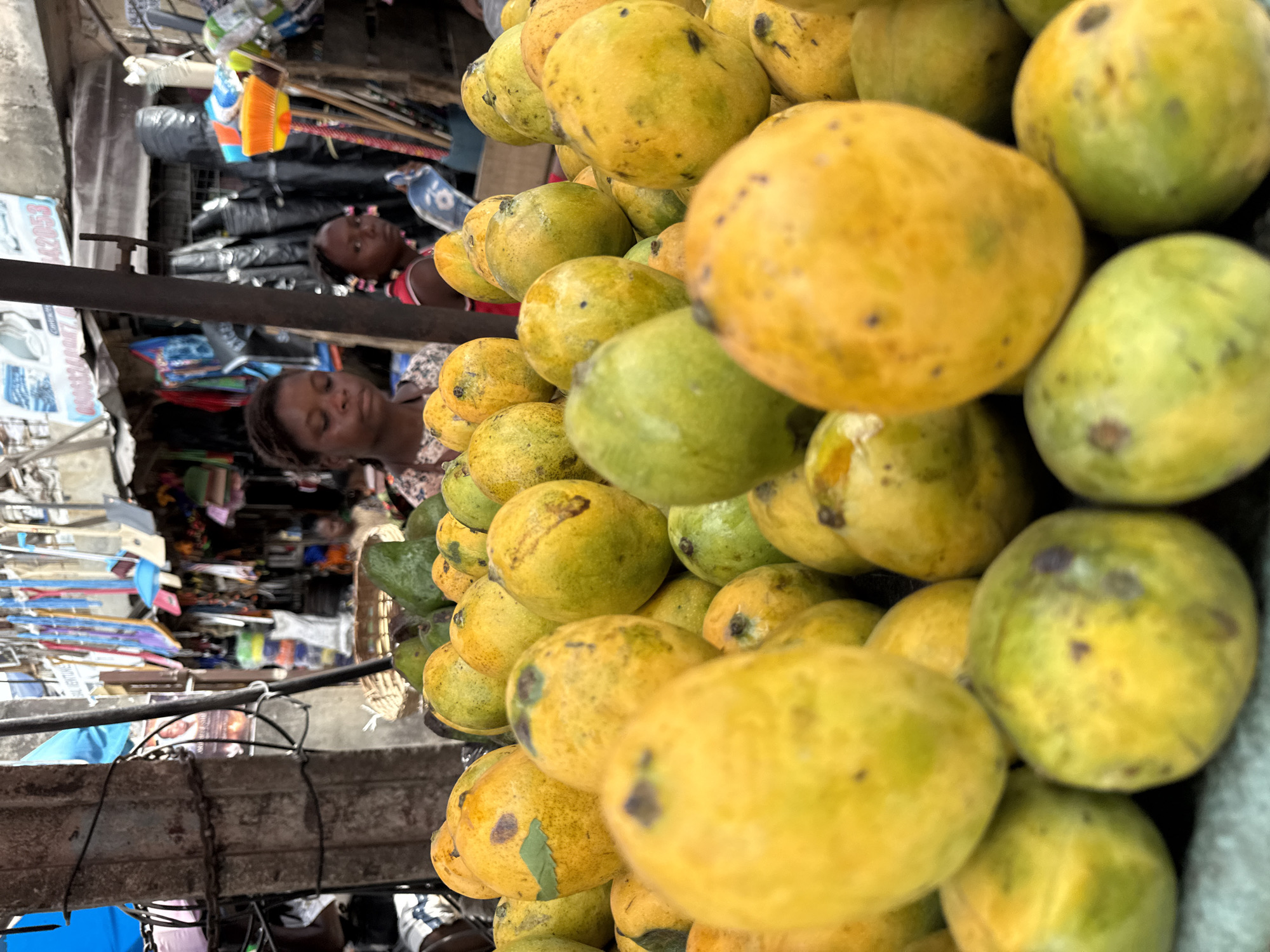 The photograph depicts a vendor selling mangoes in Lagos, Nigeria. The foreground is dominated by a large pile of unripe and ripe mangoes in various shades of green and yellow. In the background, two African women appear to be the vendor. The environment suggests a bustling marketplace with various wares and goods on display. The overall image captures the everyday scene of street vending in Lagos.