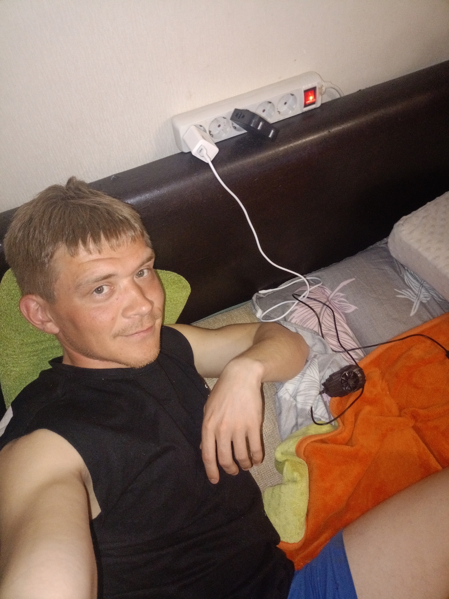 The image depicts a male individual reclining indoors in the city of Chaikovsky, Russia. The subject, an adult male with short light brown hair and light facial stubble, wears a black sleeveless t-shirt and blue shorts. He is positioned on a bed or couch, resting his head on a green textured cushion. His left arm is bent, and he looks directly at the camera with a slight smile. An orange fuzzy blanket covers a portion of his lower body, layered over a grey pillow with a subtle floral pattern. To his right, a plain light-colored pillow is visible. Above the headboard, a white power strip with a red illuminated switch is mounted on a light-colored wall. A white USB wall adapter and a black USB adapter are plugged into the power strip. A white cable extends from the white adapter, and multiple black cables connect to a small black device resembling a portable fan, positioned on the orange blanket.