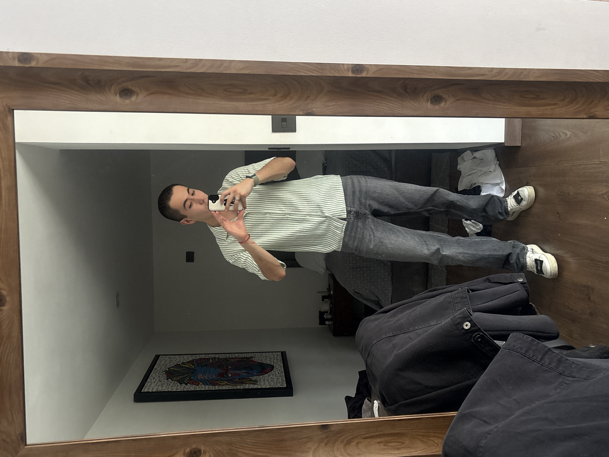 A man is taking a selfie in a mirror. He is wearing a striped short-sleeved shirt, grey jeans, and white sneakers. He is holding a smartphone in his right hand. The mirror has a wooden frame. In the background, there is a painting on the wall, a bed, and a pile of clothes. The floor is made of wood. The location is possibly an apartment in San Bernardino Tlaxcalancingo, Mexico.