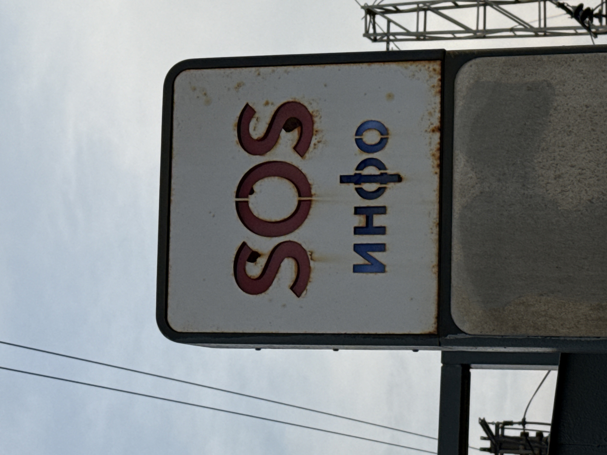 An outdoor, slightly tilted, medium shot captures a weather-beaten sign against a pale, overcast sky. The sign, with a dark frame, displays "SOS" in large, maroon, stenciled letters at the top. Below this, in blue, stenciled letters, is the word "инфо", which translates to "info" in English. The sign shows signs of rust and staining. A concrete pillar supports the sign. In the background, a tall metal structure, possibly part of a pylon or tower, is visible to the right, and overhead wires run diagonally across the sky from the upper left. The scene appears to be an outdoor informational kiosk, possibly located near a transportation hub in Moscow, Russia, given the Cyrillic lettering.