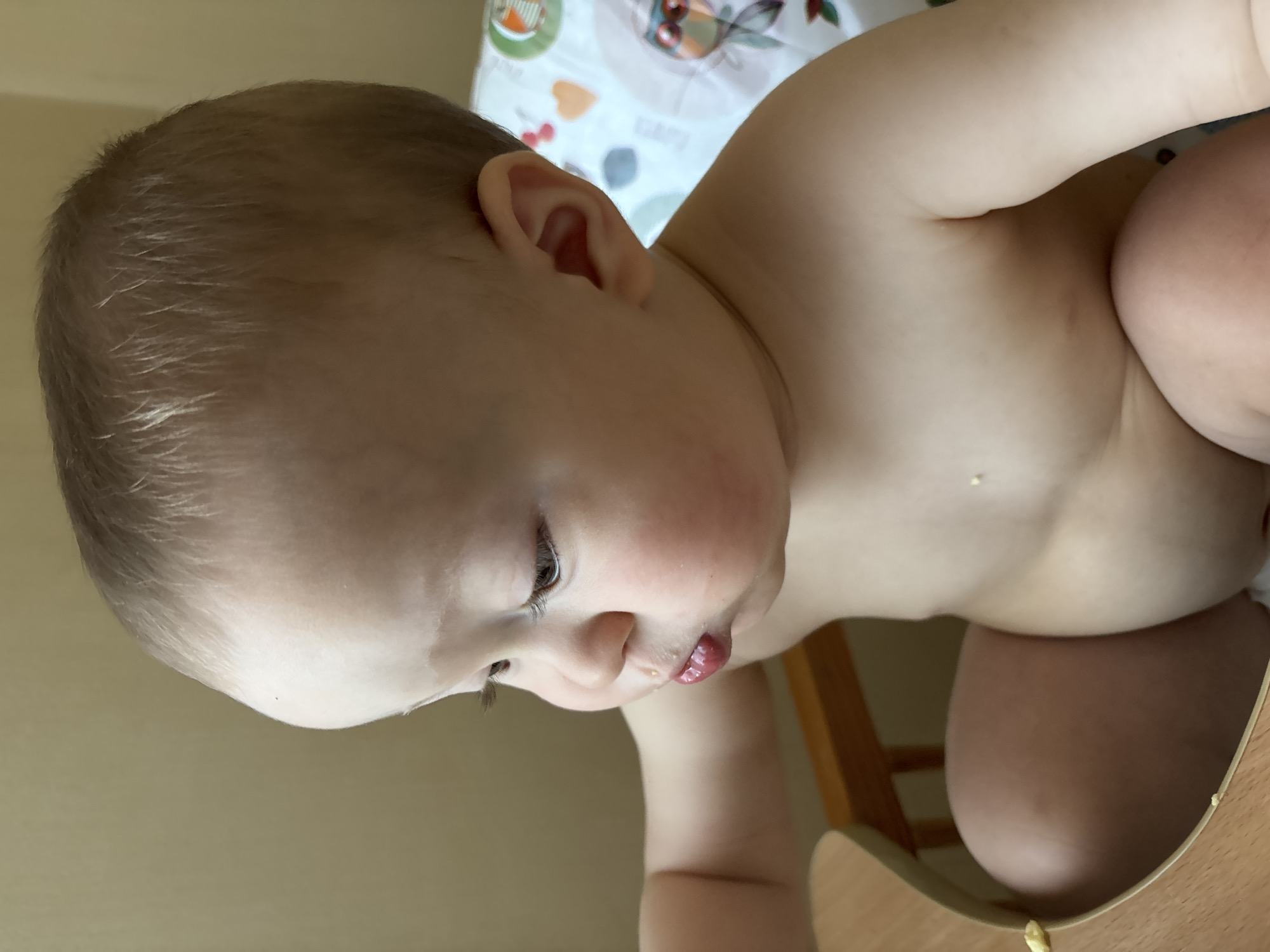 The image shows a close-up of a baby sitting in a high chair, looking down and to the left. The baby is facing to the right of the frame, with its head tilted downwards. The baby has light brown hair, fair skin, and plump cheeks. A small piece of food is visible on the baby's lower lip. The baby is wearing no clothes, exposing its torso and legs. The background is a plain, light-colored wall. A patterned bib is partially visible behind the baby's right shoulder. Part of the wooden high chair is visible in the foreground. There is no indication of the location being Naberezhnye Chelny, Russia, or any other specific city.