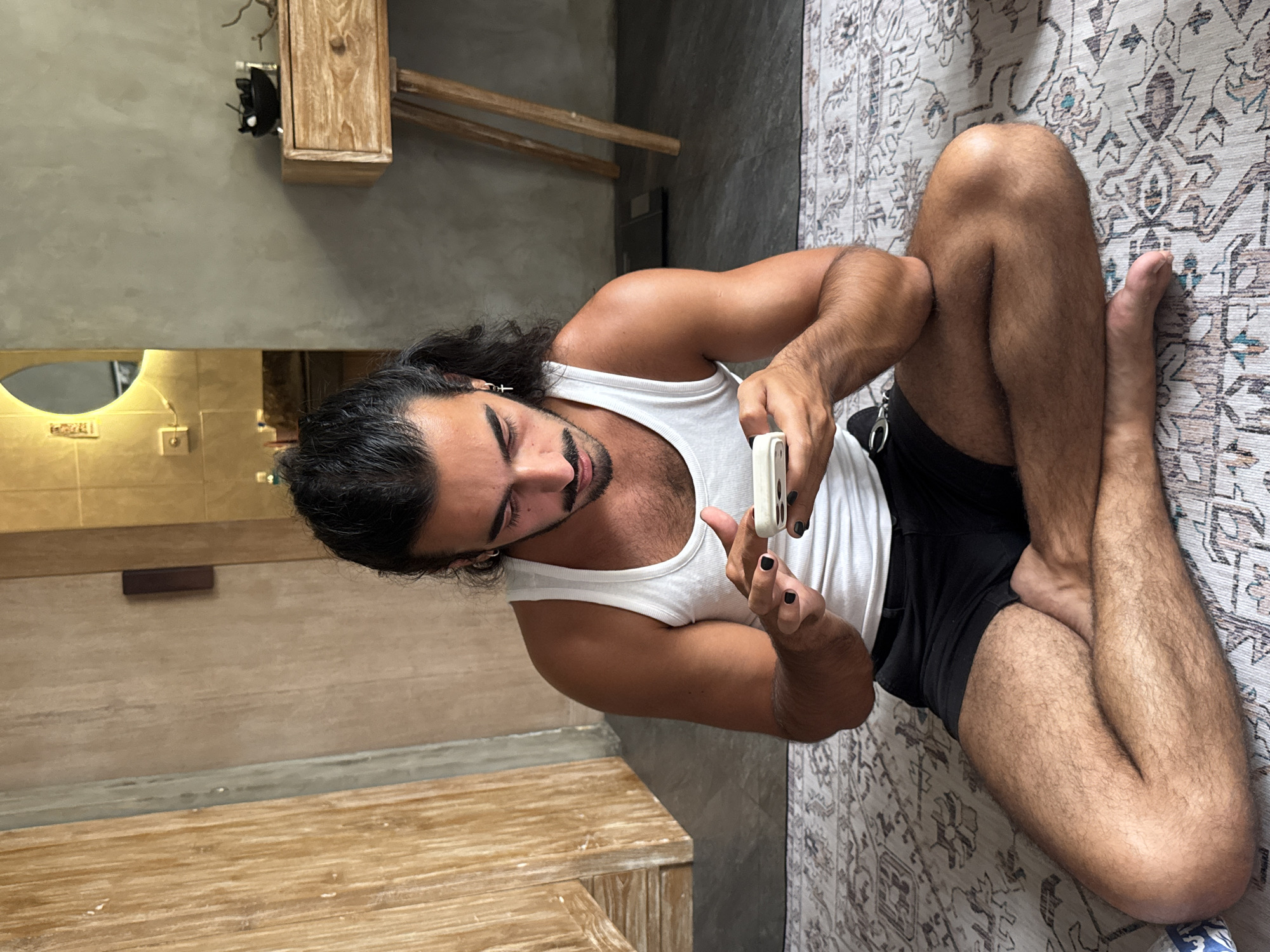 A man with long dark hair tied in a bun and a mustache is sitting cross-legged on a patterned rug. He is wearing a white tank top and black shorts. The man is looking down at his white smartphone, which he is holding in his hands. His fingernails are painted black. The background shows a wooden door on the left, a wall with a mirror and light fixtures in the center, and a wooden side table with a bowl on the right. A scale is visible on the floor behind the man. The lighting suggests an indoor environment, possibly a bathroom or dressing area within a home in North Kuta, Indonesia.
