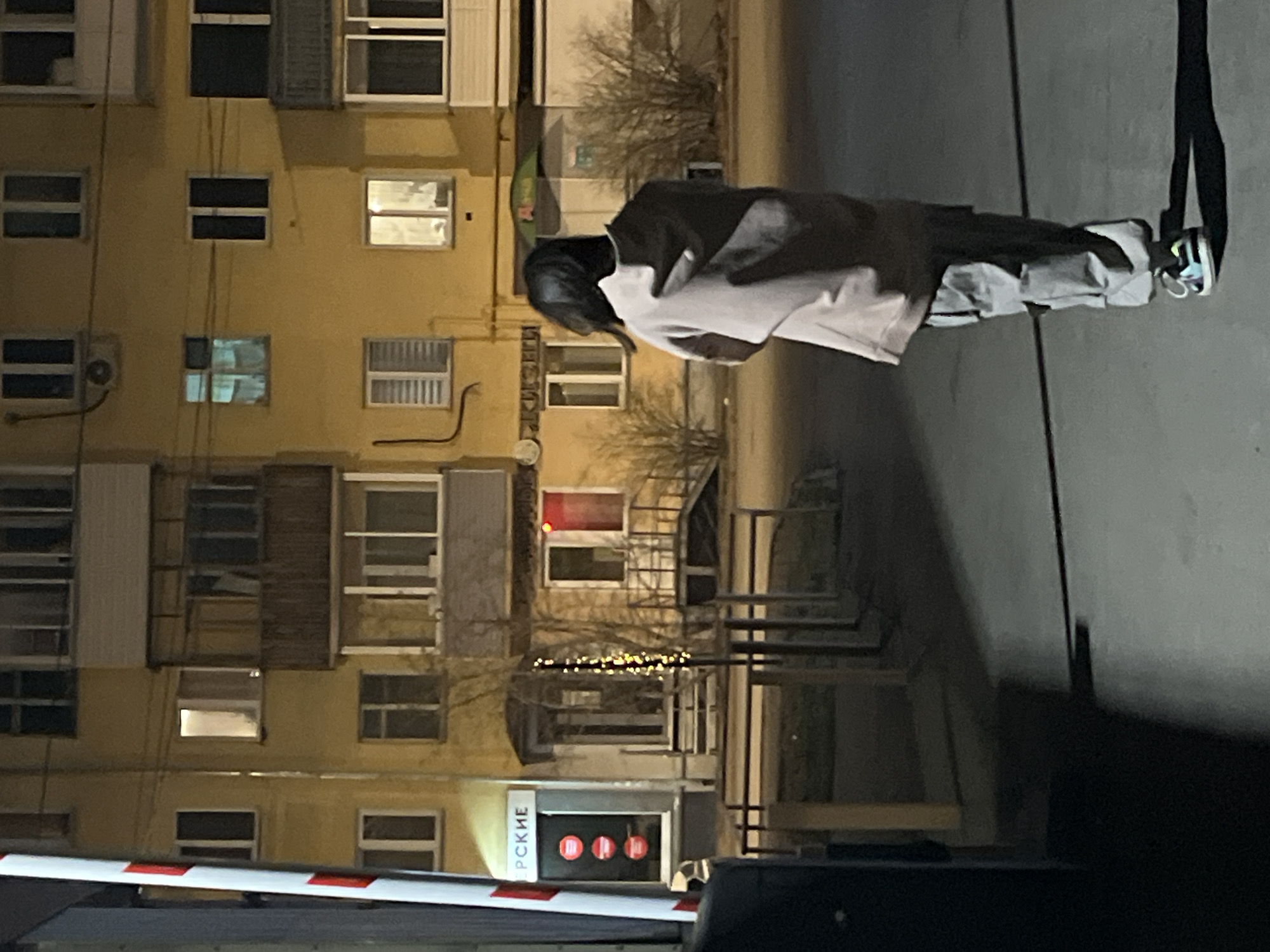 The image depicts a person standing outdoors at night, with a building in the background. The person is wearing a two-toned jacket, dark pants, and sneakers. The building has many windows, some with closed shutters. A barrier with red lights and signage in a non-English script is visible in the foreground. The location appears to be urban.
