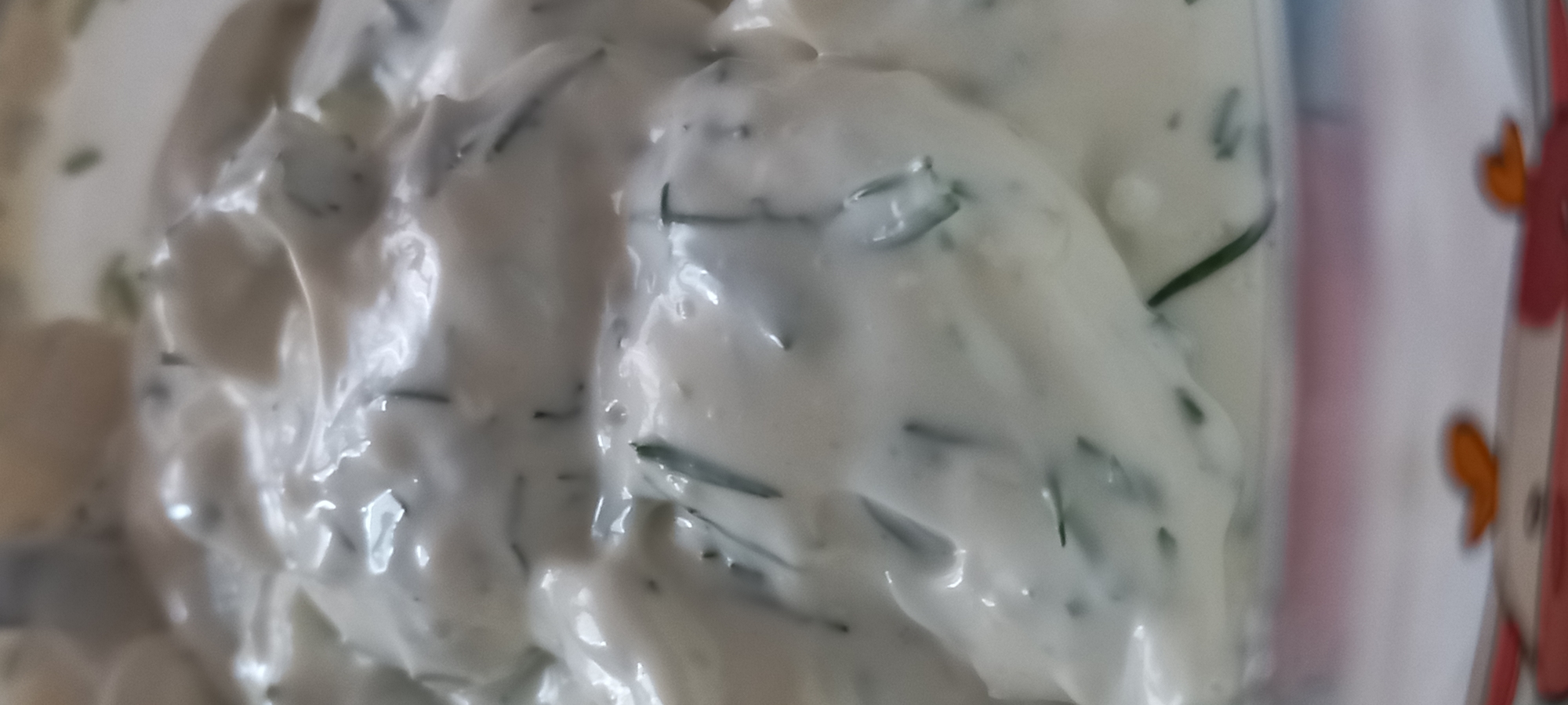 The image provides a close-up view of a thick, creamy white substance, consistent with a sauce or dip, liberally interspersed with small, dark green fragments of what appear to be fresh herbs. The surface of the substance is textured, showing soft peaks and valleys, with reflective highlights indicating a moist consistency. The bottom edge of the frame reveals a blurred, decorated surface of a container, featuring reddish and orange elements on a lighter background, possibly a patterned dish. There is no visual information within the image to determine a specific geographic location, such as the city of Kryvyi Rih, Ukraine. No individuals are present. No specific actions or interactions are depicted beyond the static presentation of the food item.