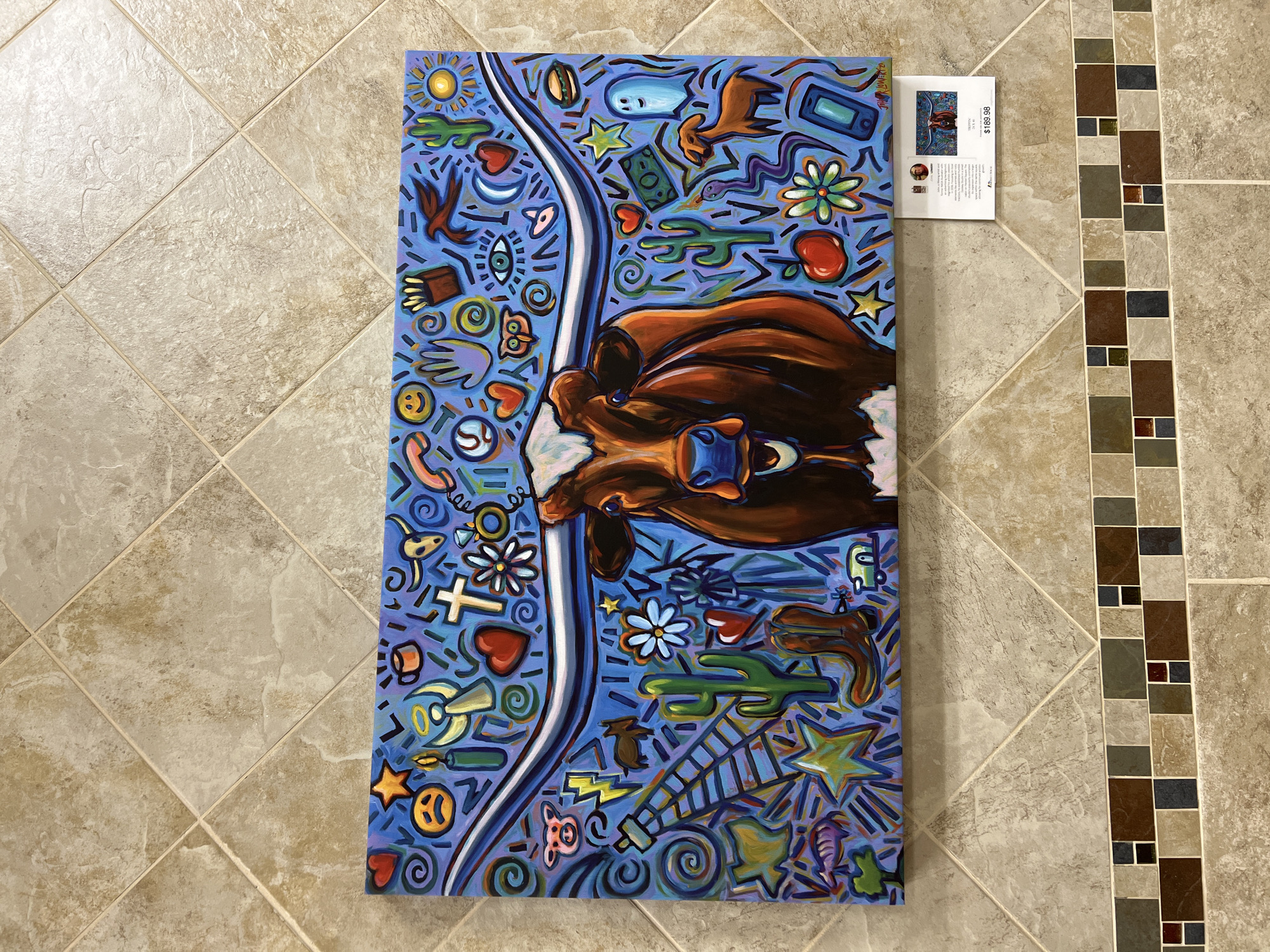 The image shows a colorful acrylic painting of a Texas Longhorn cow with large horns set against a blue background filled with various small icons and symbols. The cow is depicted from the chest up, with its head turned slightly to the viewer's right. Its eyes are wide open, and its mouth is open in a smile, revealing its teeth. The background is a vibrant blue and is adorned with a multitude of colorful, cartoonish illustrations, including a smiling sun, various hearts, stars, lightning bolts, a cowboy boot, cacti, an owl, an eye, a moon, a ghost, a hamburger, a phone, and a dollar bill, among others. The painting is framed by light brown and beige square tiles on the wall, with a decorative border of smaller, multi-colored square tiles below the painting. A small white card with text and an image of the painting is attached to the bottom right corner of the artwork, displaying a price of "$189.98". The painting is hung on a wall in what appears to be an interior space, possibly an art gallery or a shop.