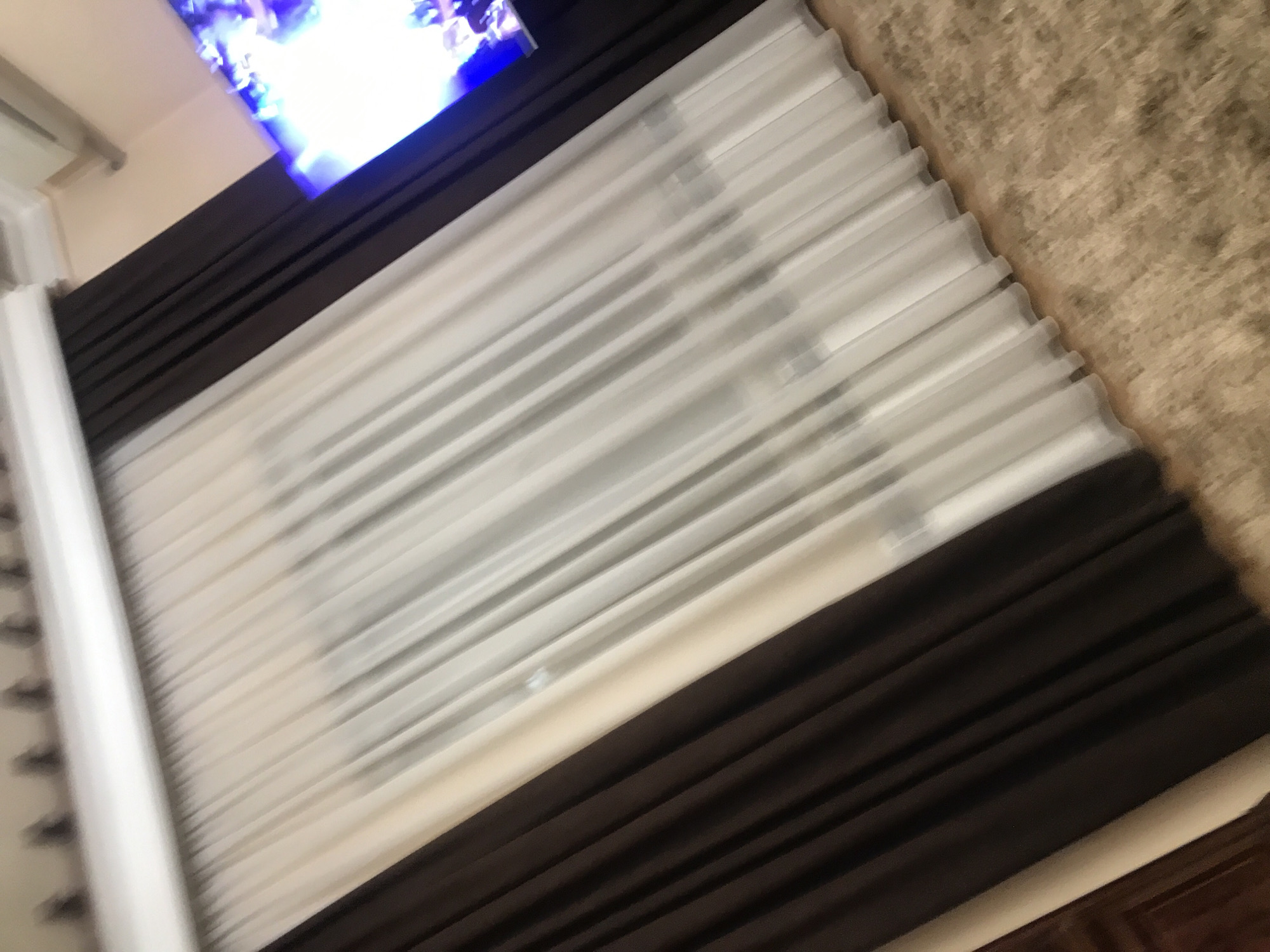 The image shows a window covered with two layers of curtains, a light colored sheer curtain layer overlaid with a dark brown curtain layer. The curtains appear to be in a residential setting, possibly in Yerevan, Armenia. A portion of a television screen is visible above the window, displaying bright colors. A textured surface, potentially a wall or carpet, is present to the right of the window. The image is taken at an angle.