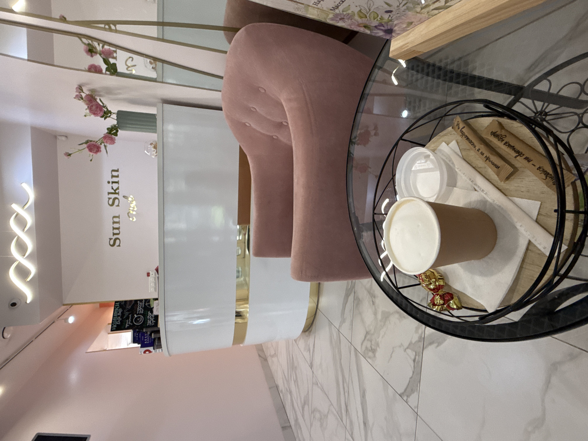 The image depicts the interior of a beauty salon. In the foreground, a black metal circular coffee table with a glass top holds a tray with a disposable coffee cup filled with a foamy beverage, a small plastic lid, two wrapped candies, and two paper sugar packets with Cyrillic text. To the right of the tray, a wooden plank leans against the table.
The background features a plush pink armchair and a reception desk with a white and gold facade. The desk has the text "Sun Skin" and "Nail" in gold lettering. A vase with pink flowers sits on the desk. A large mirror is positioned behind the desk, reflecting the room. The floor is tiled with a marble pattern. A pink wall is visible on the left side of the image, and a dark rectangle, possibly a screen, is mounted on it. A menu board with handwritten text is placed on the reception desk. The lighting appears to be soft and ambient.
