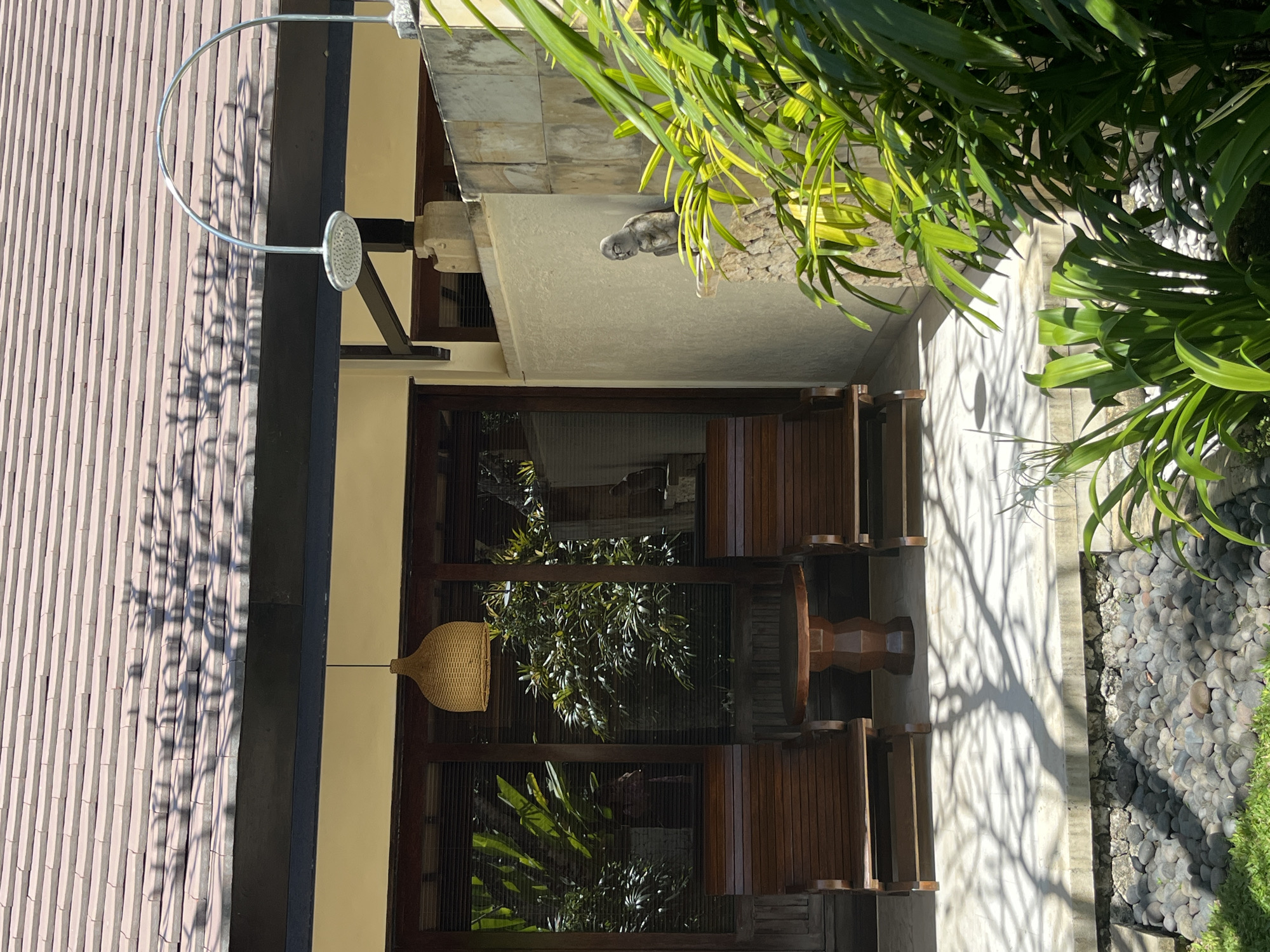 The image depicts an outdoor seating area at what appears to be a resort or villa in Kuta Selatan, Indonesia. The scene is set under a tiled roof with shadows from overhead foliage cast across the ground. In the foreground, lush green plants are visible, some of which are in focus.
The seating area features two wooden lounge chairs flanking a small, round wooden table. Behind the chairs, a glass-paned door or window leads into a building, revealing interior lighting and more greenery. An overhead lamp with a woven shade hangs above the seating arrangement.
To the right of the seating area, a stone wall is prominent, with a modern outdoor shower head arching over it. Perched on the stone wall is a small, gray stone statue of a crouching figure with distinct facial features. The ground in front of the seating area is a mixture of concrete, small dark pebbles, and some green grass. The overall atmosphere suggests a tranquil, tropical setting designed for relaxation.