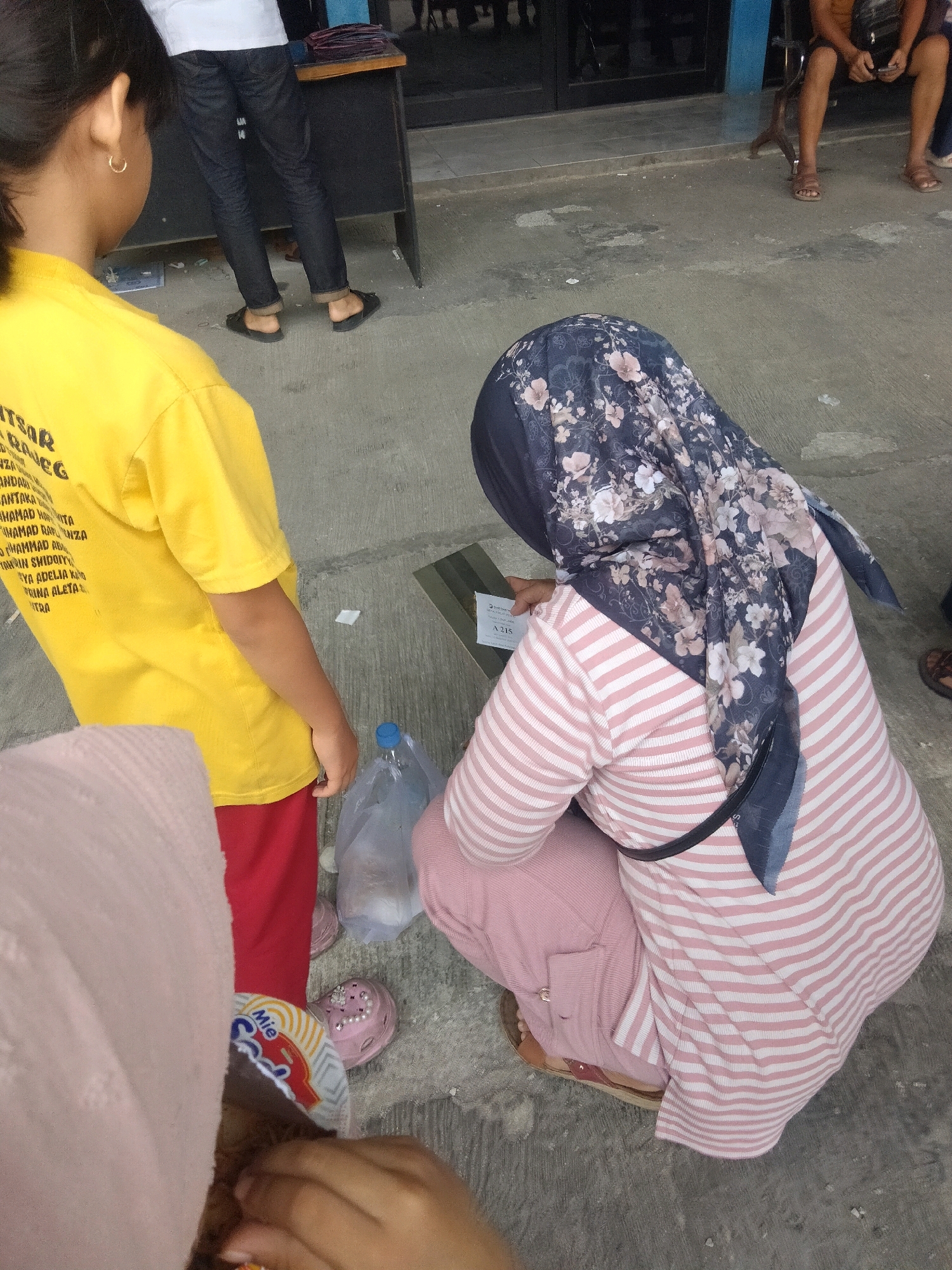 In Sukaasih, Indonesia, a young girl in a yellow t-shirt stands next to a person wearing a pink and white striped long-sleeved shirt and a floral hijab. The person in the hijab is squatting and holding a white slip of paper. A plastic water bottle and a clear plastic bag containing an object are on the ground near them. The ground is concrete with some wear and tear. In the background, another person in a white t-shirt and black jeans is standing next to a dark desk. The scene appears to be outdoors, possibly in a public area.