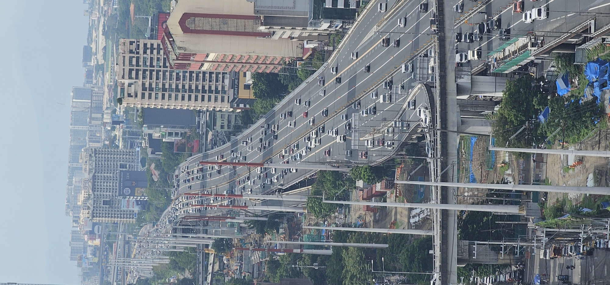 This is a vertical, high-angle aerial photograph taken in Makati, Philippines, depicting a multi-level elevated highway system with moderate to heavy traffic. The highway is characterized by concrete pillars and barriers, with multiple lanes of vehicles including cars, vans, and some trucks. Some sections of the highway appear to be under construction or undergoing maintenance, with visible scaffolding, construction materials, and temporary barriers.
In the background, a dense urban landscape unfolds, featuring numerous high-rise buildings of varying architectural styles and heights, indicating a commercial and residential district. The buildings are a mix of modern skyscrapers and older structures, some with visible advertisements and signage. A hazy sky suggests a warm and possibly humid climate.
The foreground shows a lower level of infrastructure, possibly a road or another part of the highway system, with more traffic and construction activity. Below the elevated highway, there are areas with greenery, trees, and some temporary shelters or makeshift structures, suggesting the presence of informal settlements or work areas associated with the construction. The overall scene conveys a sense of a bustling, dynamic urban environment with ongoing development and significant vehicular movement.