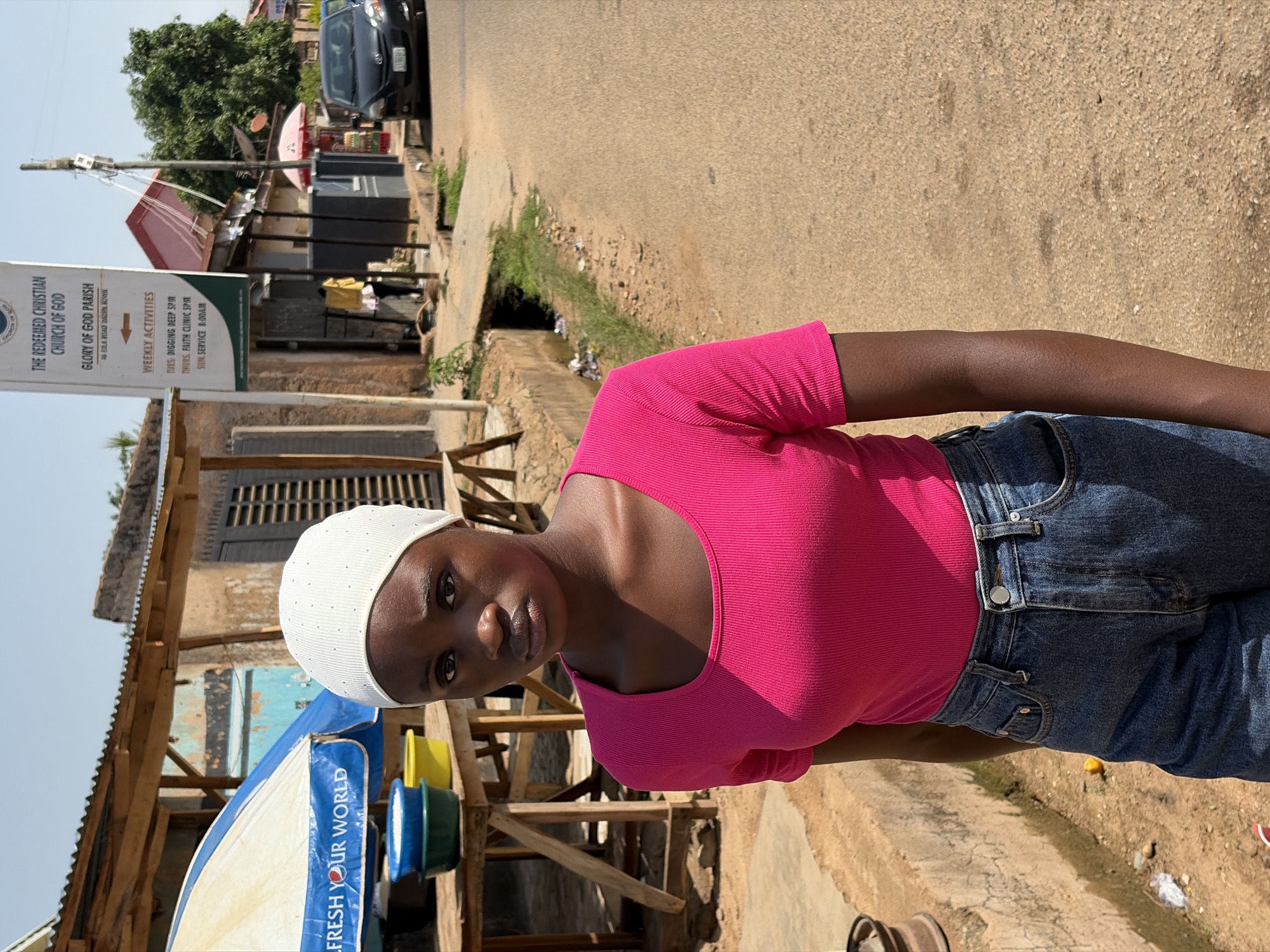 A young woman with dark skin, wearing a white cap, a pink top, and jeans, stands in front of a building and a street. The sign for "THE REDEEMED CHRISTIAN CHURCH OF GOD, GLORY OF GOD PARISH, 16 ELA ROAD DADIN KOWA" is visible. A partly visible car is on the street. The location is the city of Jos, Nigeria.
