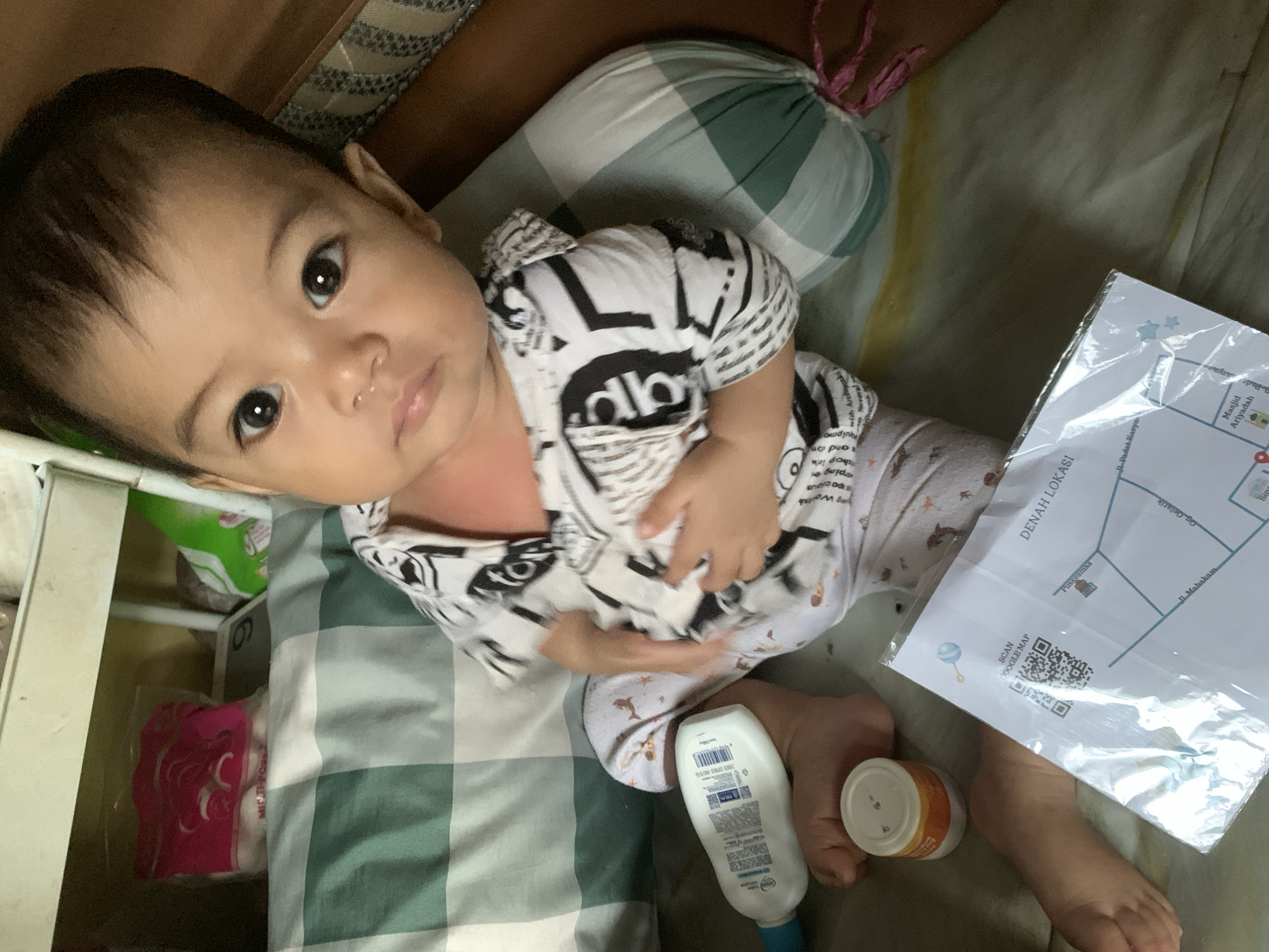 The image shows a baby lying on a green and white checkered surface, likely a bed or play area. The baby is wearing a black and white patterned shirt and light-colored pants. Next to the baby is a tube of lotion, a small jar, and a document titled "DENAH LOKASI" with a map and a QR code labeled "SCAN GOOGLE MAP." The map includes place names such as Jl. Padat Karya, Masjid Ariyadah, Jl. Mahakam, Gg. Gelatik, and Puskesmas. Based on these locations, the scene is in Sungai Meriam, Indonesia.
