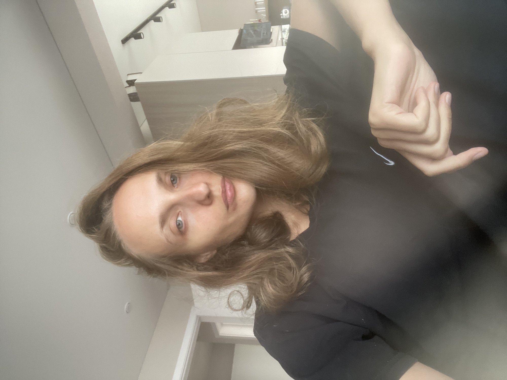 The image is a close-up, waist-up selfie of a woman with light brown, medium-length hair, taken indoors. She is wearing a black t-shirt with a small white Nike logo. Her gaze is directed towards the camera, and her mouth is slightly pursed. Her right arm and hand are visible in the foreground, with her fingers curled. The background suggests an indoor domestic setting, possibly an apartment, with furniture and lighting fixtures visible. The specific location of Подшивалово, Russia is not discernible from the image itself.
