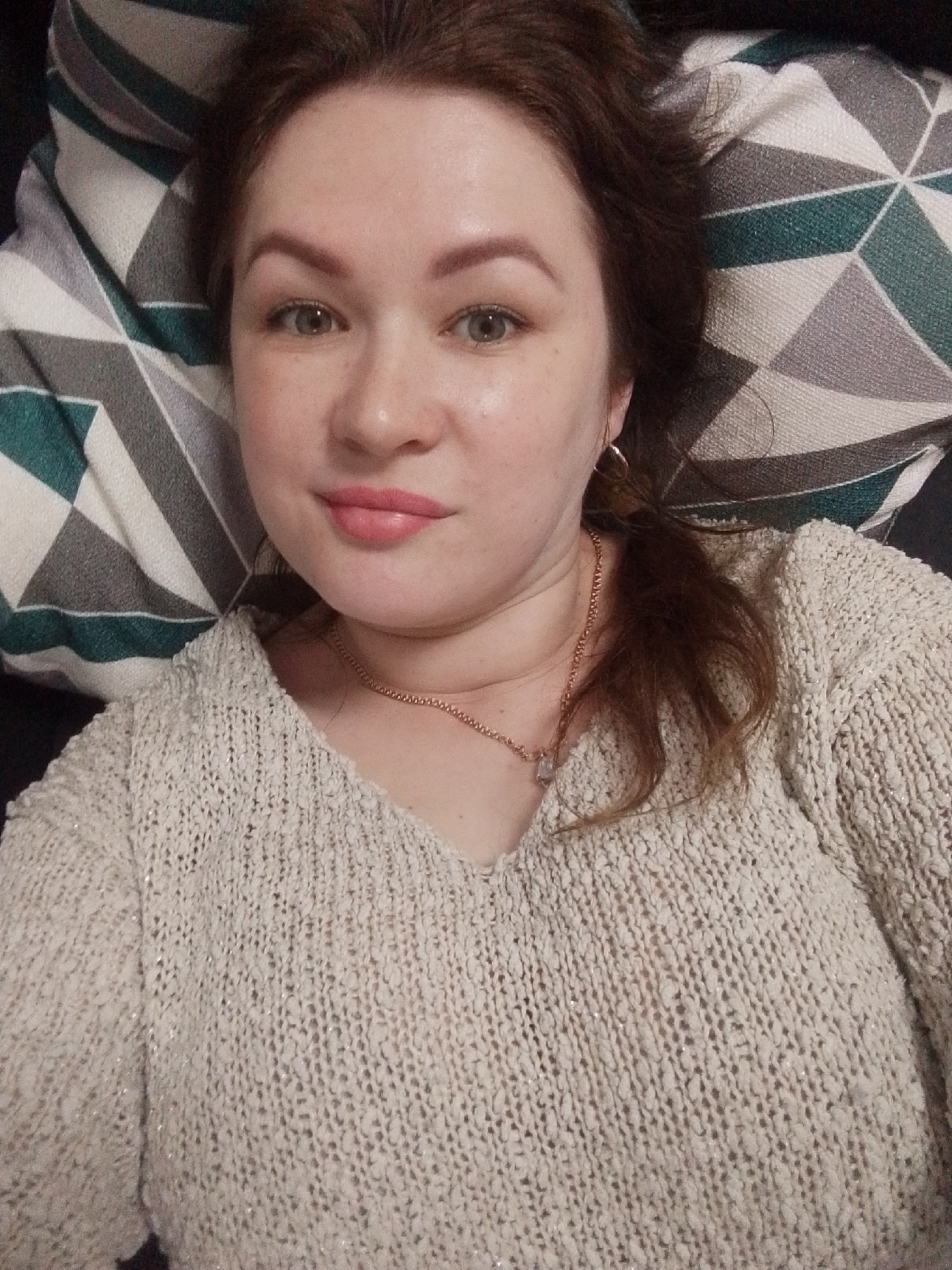 An adult woman is depicted in a close-up, head-and-shoulders selfie, positioned as if lying down or reclining, and looking directly at the camera. She has light skin, brown hair, and light-colored eyes, with a neutral to slightly smiling expression and pink lipstick on her lips. Her left ear, visible on the right side of the image, features a small, simple hoop earring.
She is wearing a V-neck, light-colored (possibly beige or off-white) knitted sweater that appears to have a textured, loose weave with subtle, shimmering threads. A delicate gold-toned chain necklace with a small, light-colored pendant rests on her chest, visible above the sweater's neckline.
Her head is resting on a decorative pillow featuring a distinct geometric pattern. This pattern comprises tessellating shapes—likely hexagons or diamonds—in shades of grey, white, and a dark teal or green. A darker, indistinct background is visible behind the pillow, suggesting an indoor setting such as a home environment. The activity is a self-portrait, and the woman appears calm and relaxed. No text is visible in the image. The provided location context is Chaikovsky, Russia.