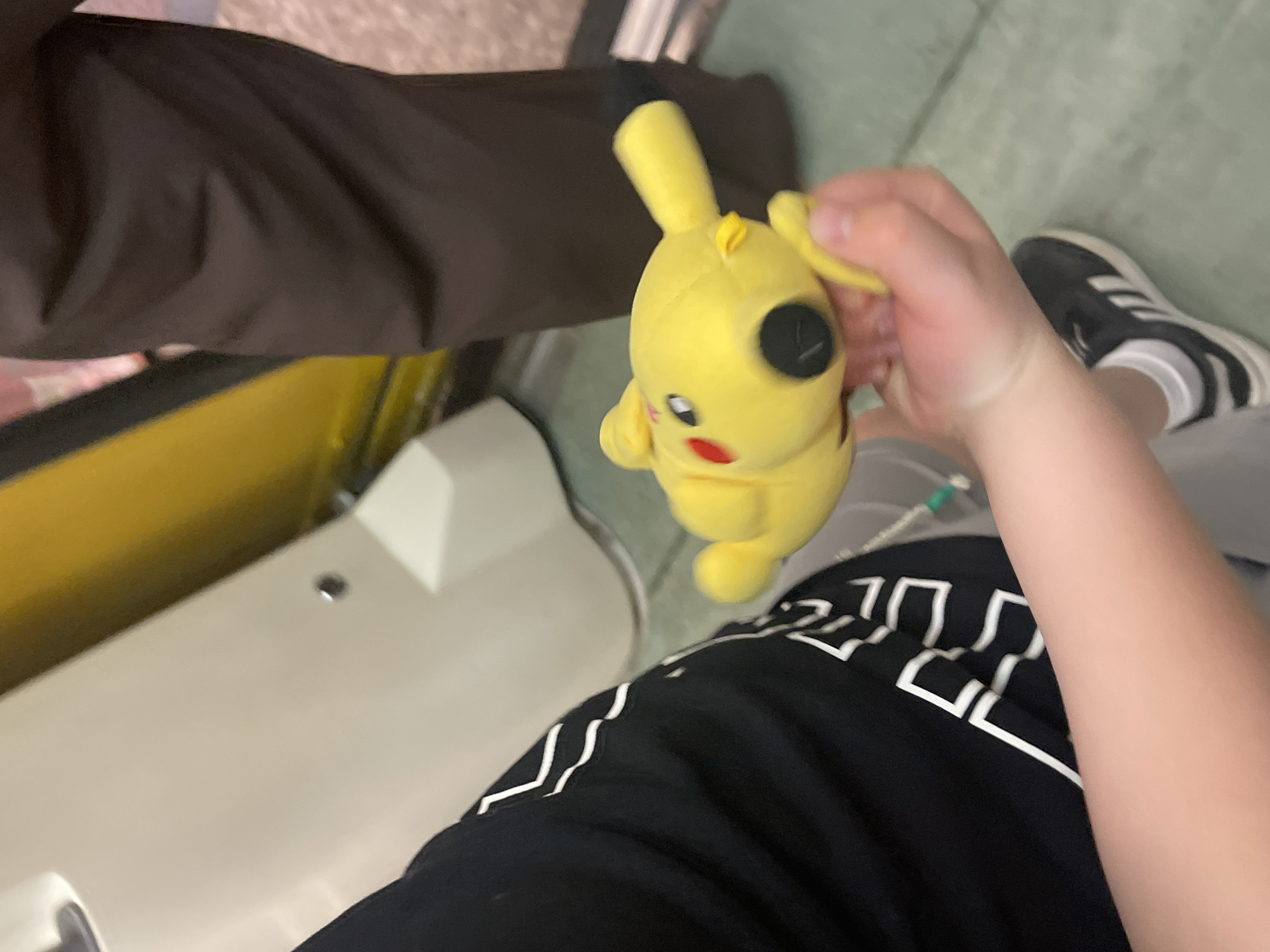 The image depicts the interior of a Moscow Metro train, identifiable by the distinctive bright yellow vertical handrail on the right side of the frame, alongside light-colored wall panels and flooring. In the foreground, a person, likely a child or young individual, is partially visible, holding a yellow Pikachu plush toy horizontally in their right hand. This person wears a black short-sleeved t-shirt with white angular patterns on the left sleeve and side, white athletic socks, and a black Adidas sneaker with three white stripes on the foot. To the right of the person holding the plush, another individual is partially visible from the lower torso down, wearing dark brown trousers. The flooring is a light greenish-grey, and various light beige or off-white structural elements of the train interior are visible in the background and to the left. A small silver stud or bolt is present on a beige panel in the upper left quadrant.